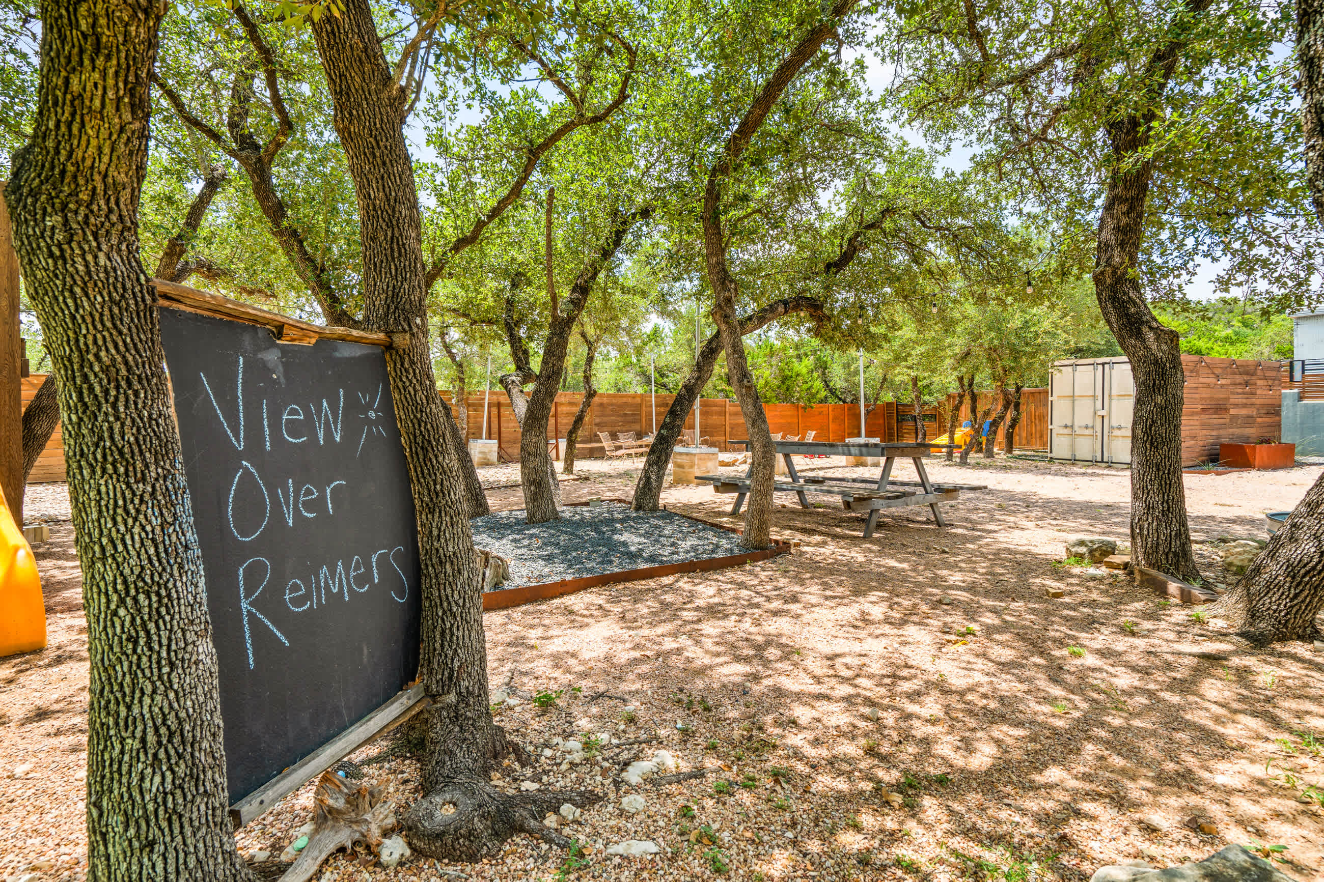 Shaded outdoor area with a rustic chalkboard sign reading "View Over Reimers" leaning against a tree. A wooden picnic table and benches are visible on a gravel surface, surrounded by mature trees.