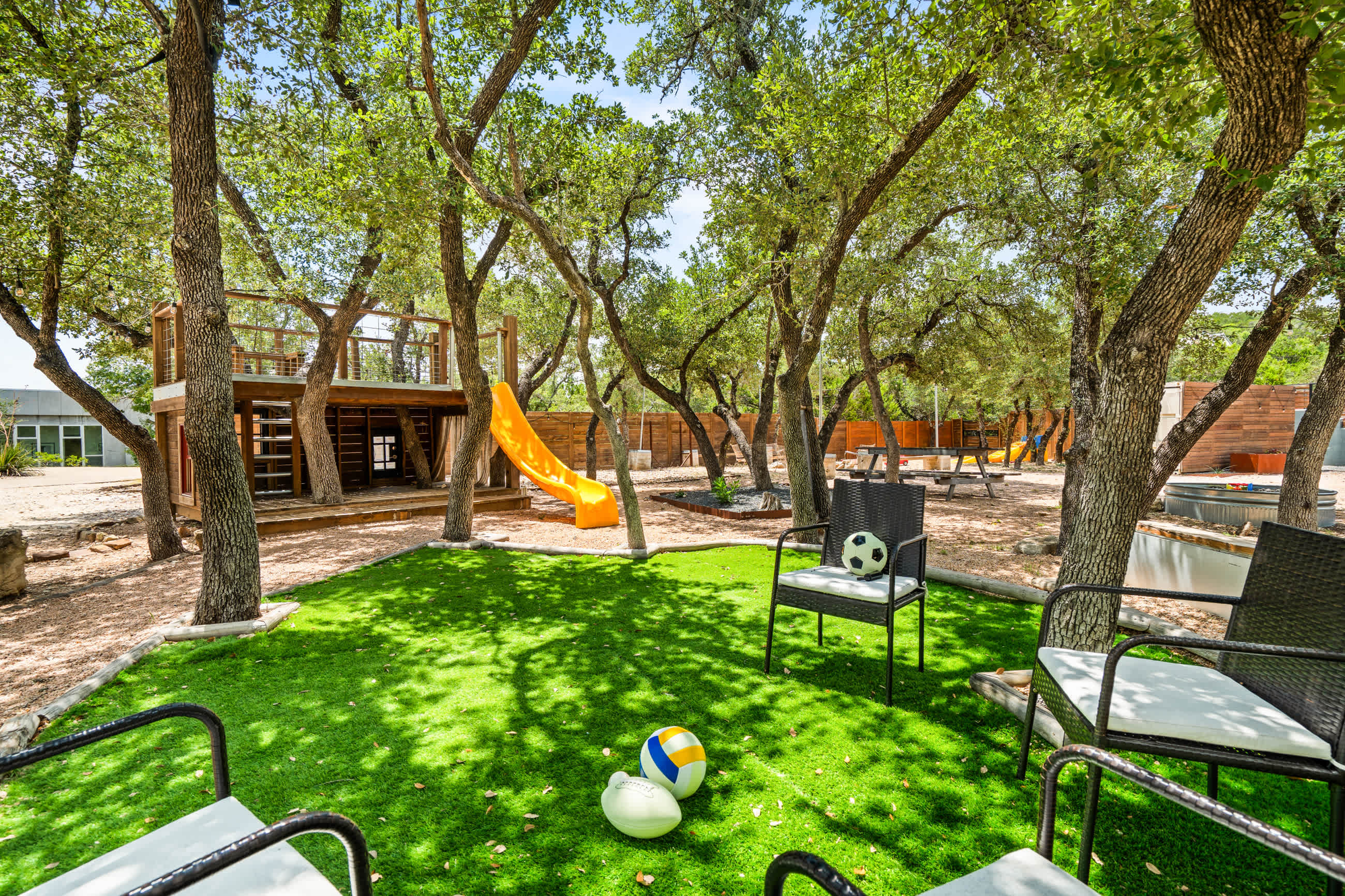 Lush artificial turf area with black outdoor chairs and a soccer ball and volleyballs, perfect for games. A wooden play structure with an orange slide is visible in the background, surrounded by mature trees.