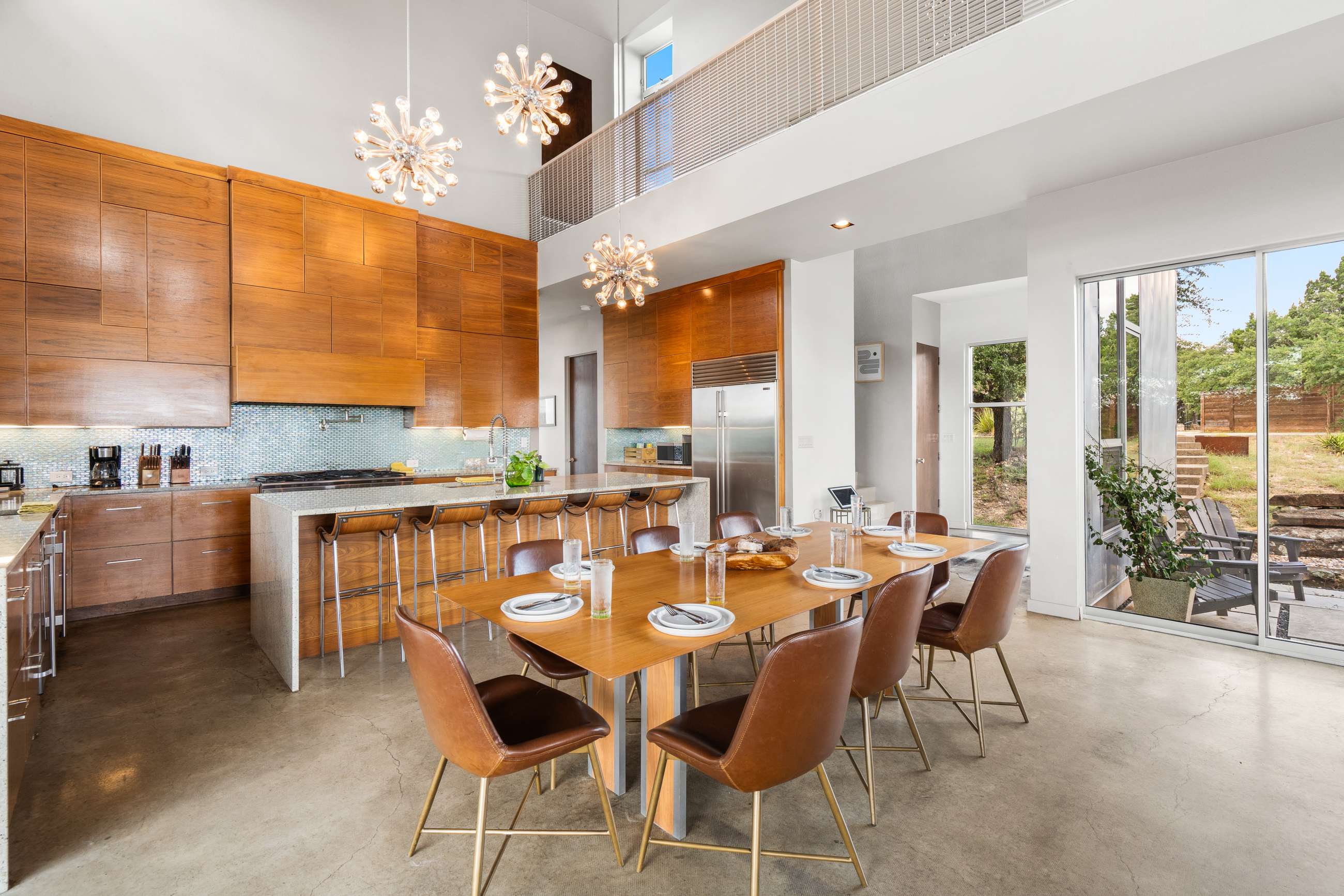 An expansive open-plan kitchen and dining space with a long wooden dining table set for eight, surrounded by brown leather chairs. The kitchen features wood cabinetry, a large island with bar seating, and a light blue tiled backsplash, all under high ceilings with modern chandeliers.