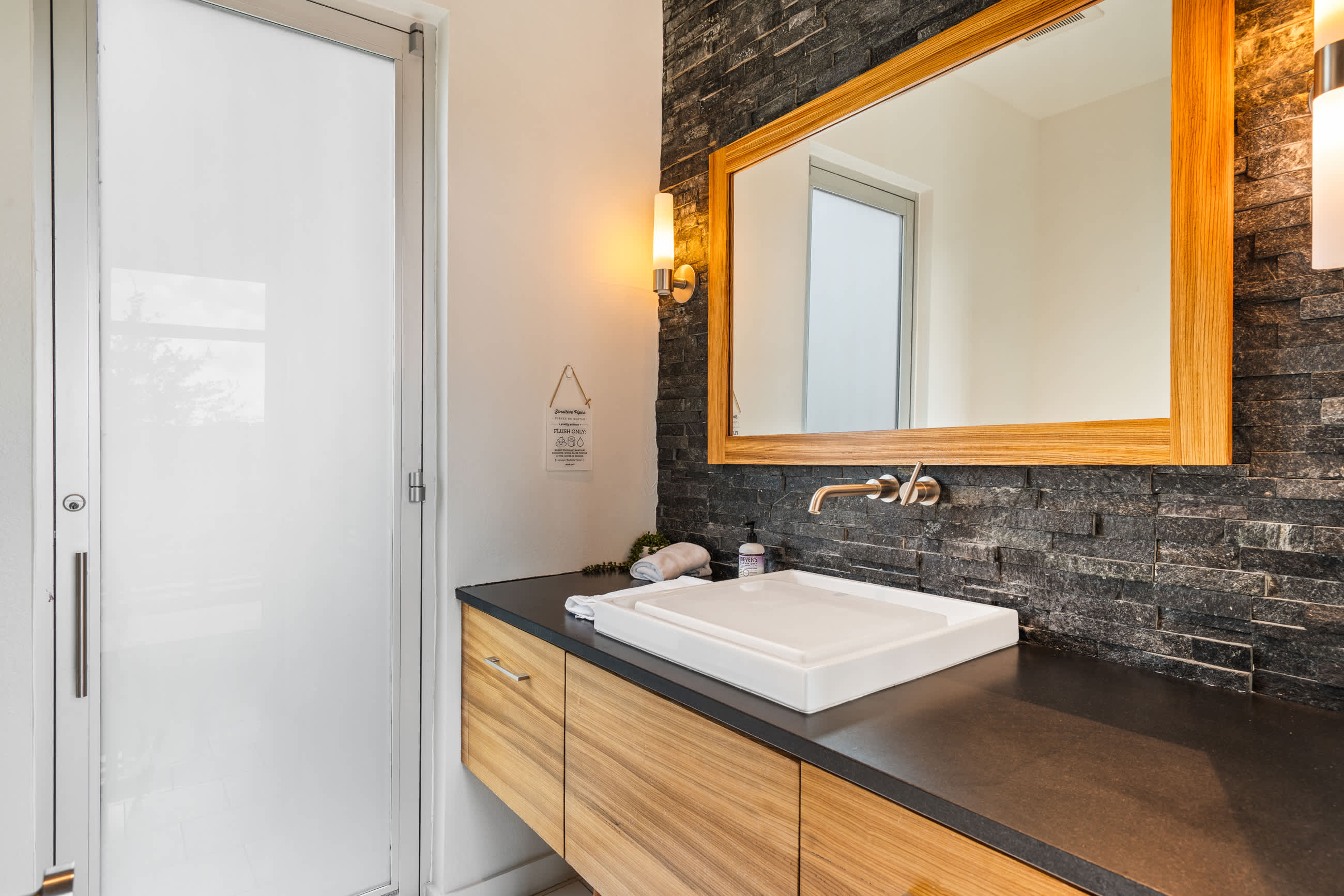 Modern bathroom featuring a white rectangular vessel sink on a dark countertop, a wooden vanity, and a large mirror with a wooden frame against a dark stacked stone accent wall. A frosted glass door is visible on the left.