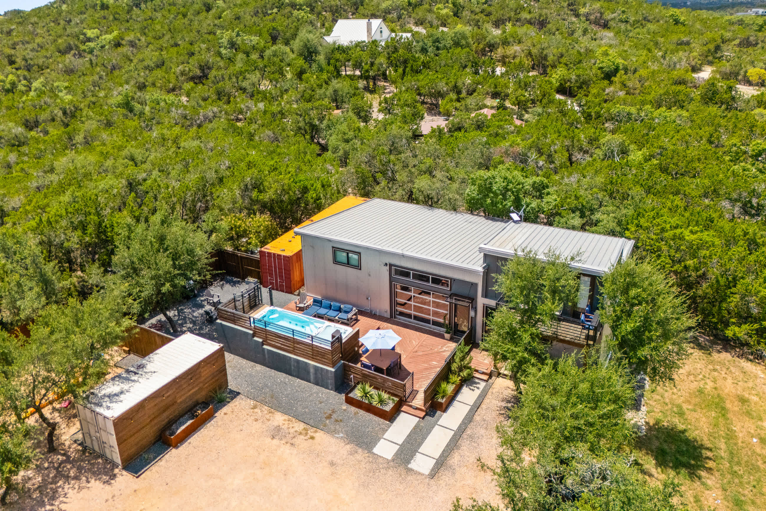 Aerial view of a modern barndominium-style structure with a metal roof, featuring an outdoor swimming pool and a wooden deck with seating. The property is surrounded by natural landscaping and gravel driveways.