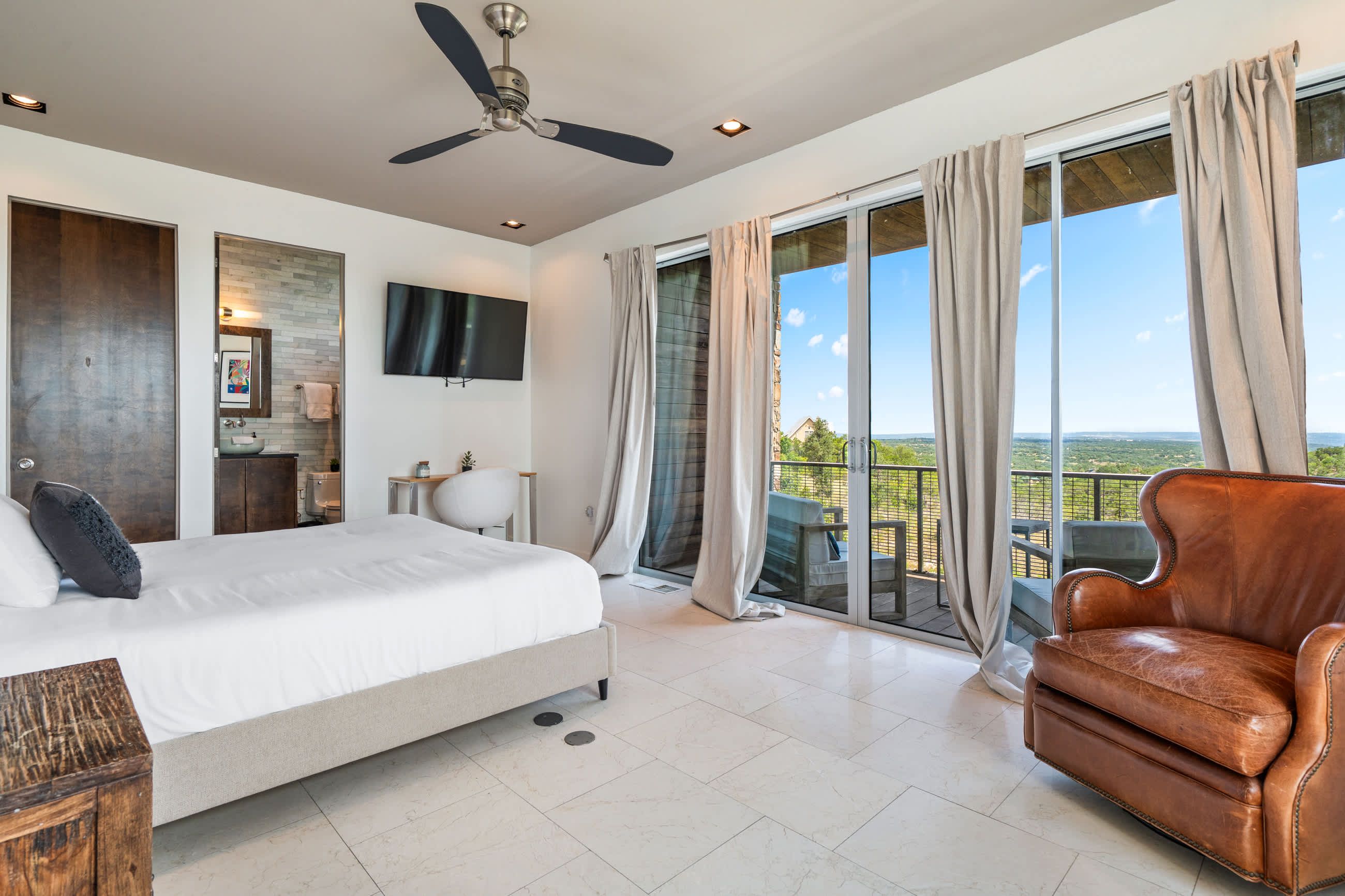 A modern bedroom with a white bed, a wall-mounted TV, a small desk, and expansive sliding glass doors opening to a balcony with scenic outdoor views. A leather armchair sits in the corner.