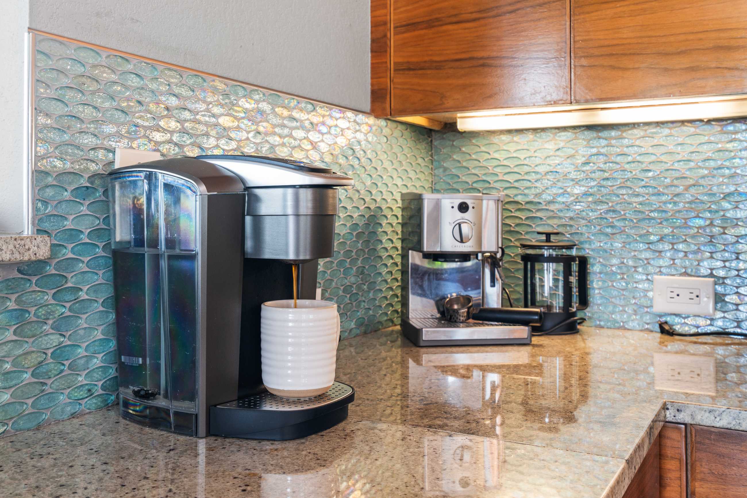 A dedicated coffee station on a granite countertop, featuring a single-serve coffee maker brewing into a white mug, an espresso machine, and a French press. The backsplash is a textured light blue mosaic tile, and wood cabinets are visible above.