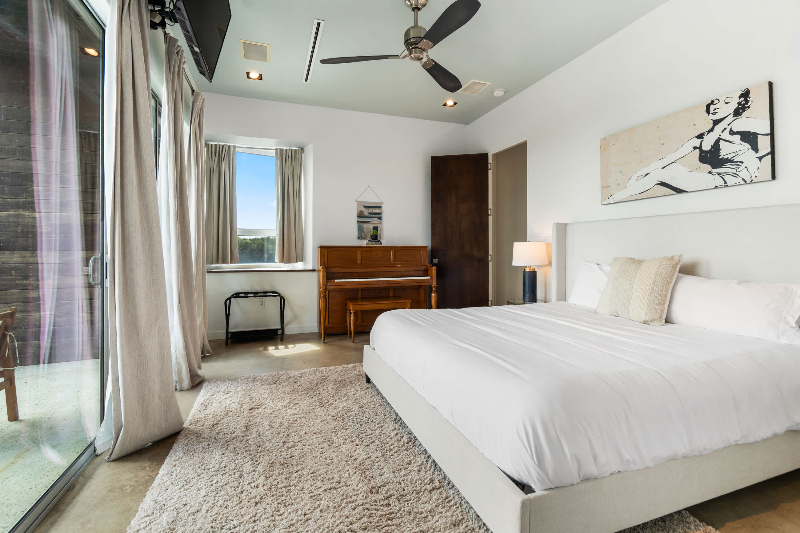 Bright bedroom featuring a white bed with a tall headboard, a wooden piano, and a large window. A modern art piece hangs above the bed, and a ceiling fan is overhead.