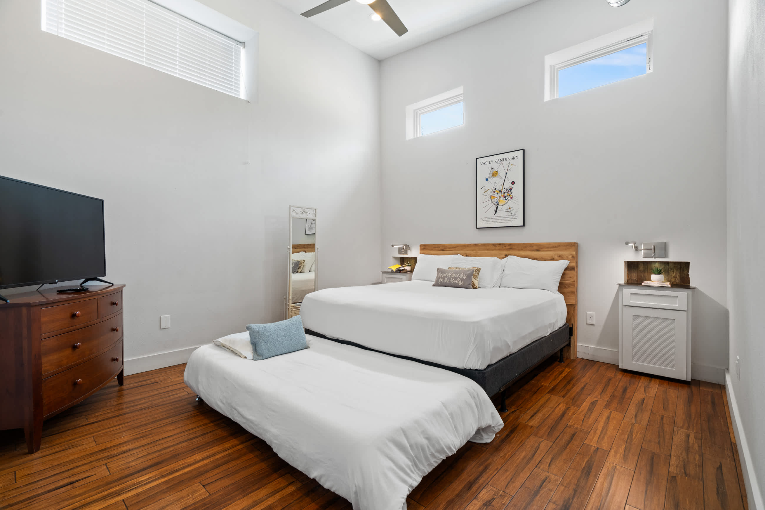 Bright bedroom with a queen bed, white bedding, a wooden headboard, and dark hardwood floors. High ceilings with small windows near the top let in natural light, and a ceiling fan is visible.