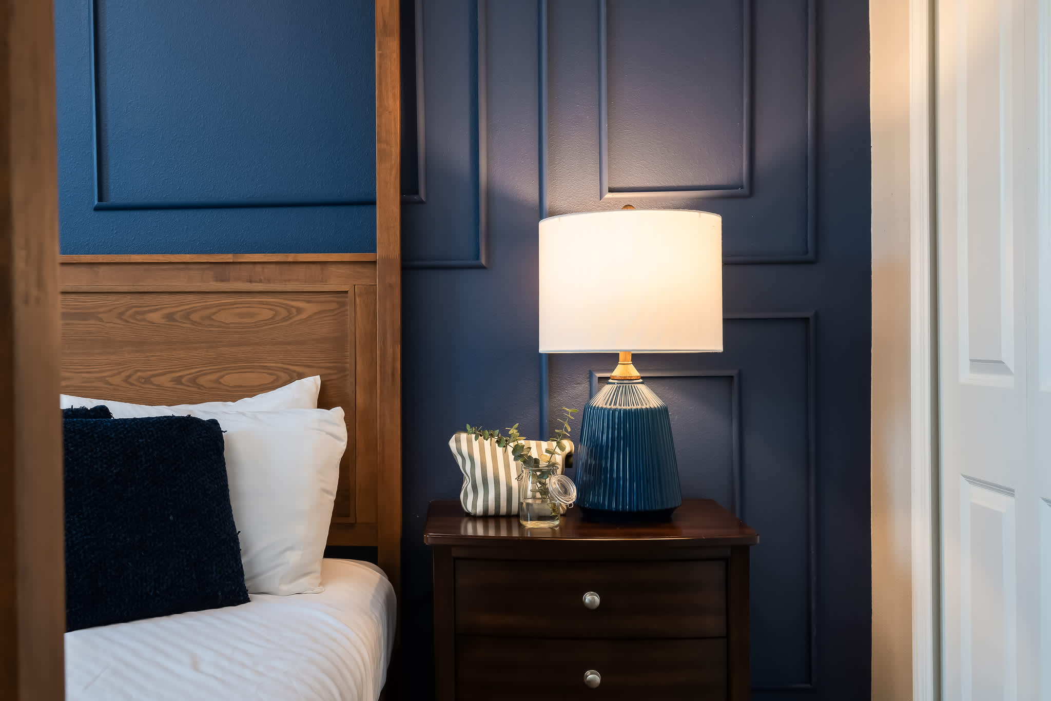 Close-up of a dark wood nightstand next to a wooden four-poster bed, featuring a blue ceramic table lamp and decorative items.
