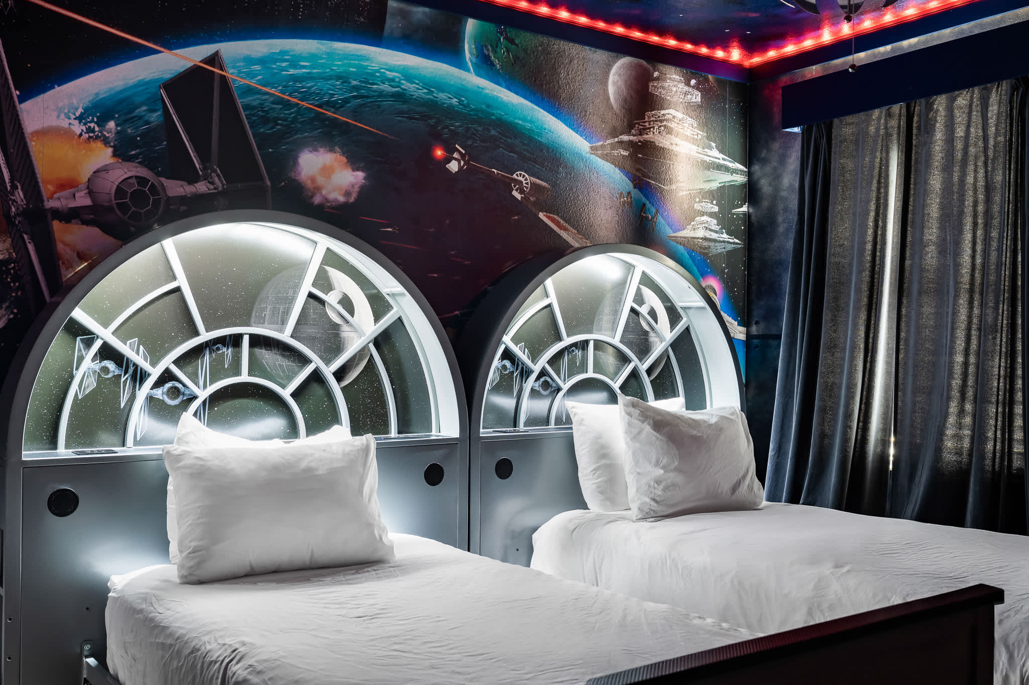 Close-up view of two twin beds in a Star Wars themed room, each with white bedding and an illuminated, circular headboard designed to look like a spaceship window, against a vibrant space mural.