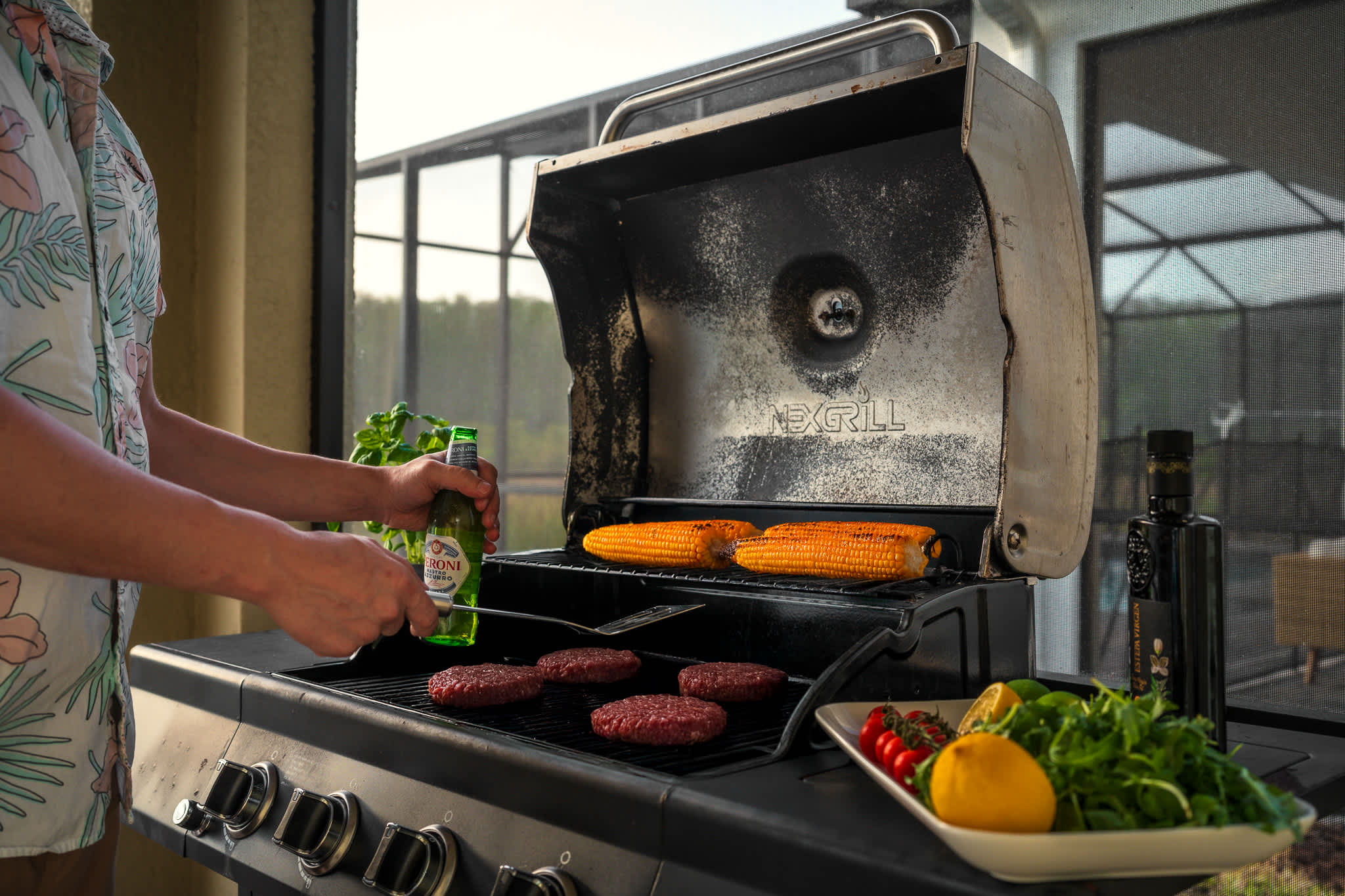 A person grills burgers and corn on a black gas grill on a patio, with a bottle of beer in hand and a platter of fresh vegetables and lemons nearby.