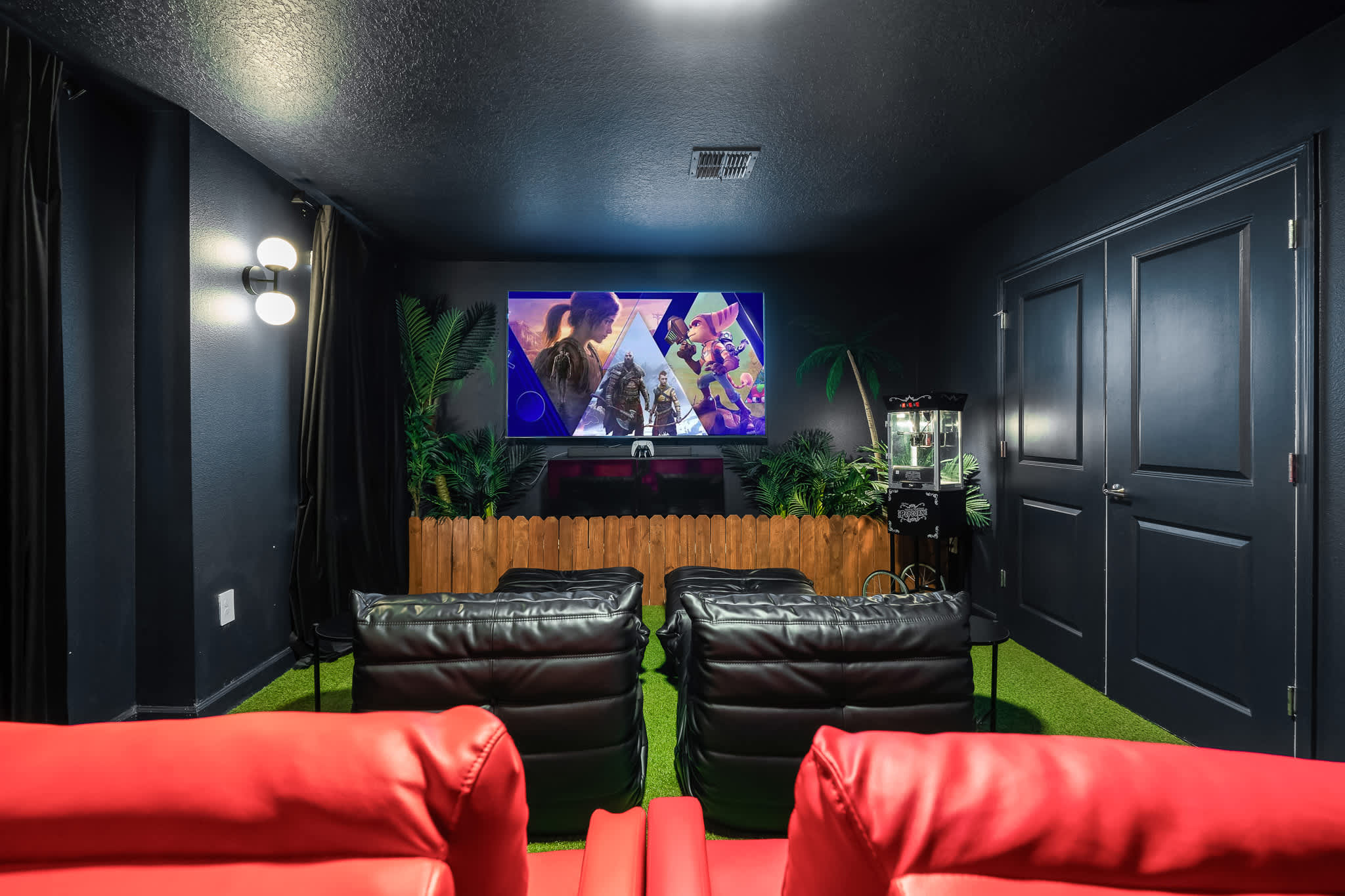 Dark-themed media room with a large screen displaying a video game, two rows of red and black recliners, and artificial turf flooring. The TV is flanked by tropical plants and a popcorn machine.