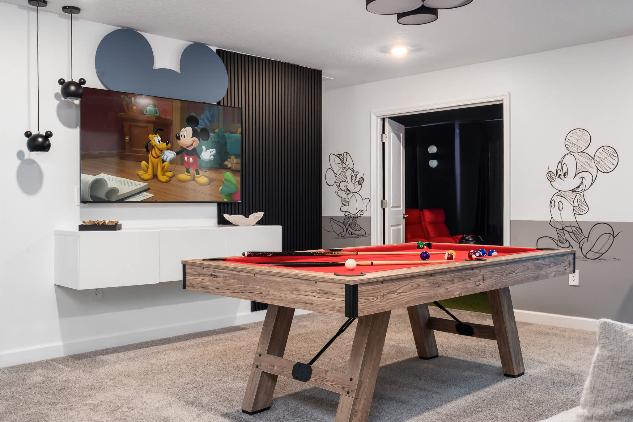 A game room showcases a red-felted pool table, a large wall-mounted TV displaying a cartoon, accented by Mickey Mouse ear decor and wall art, with a dark media room visible through an open doorway.