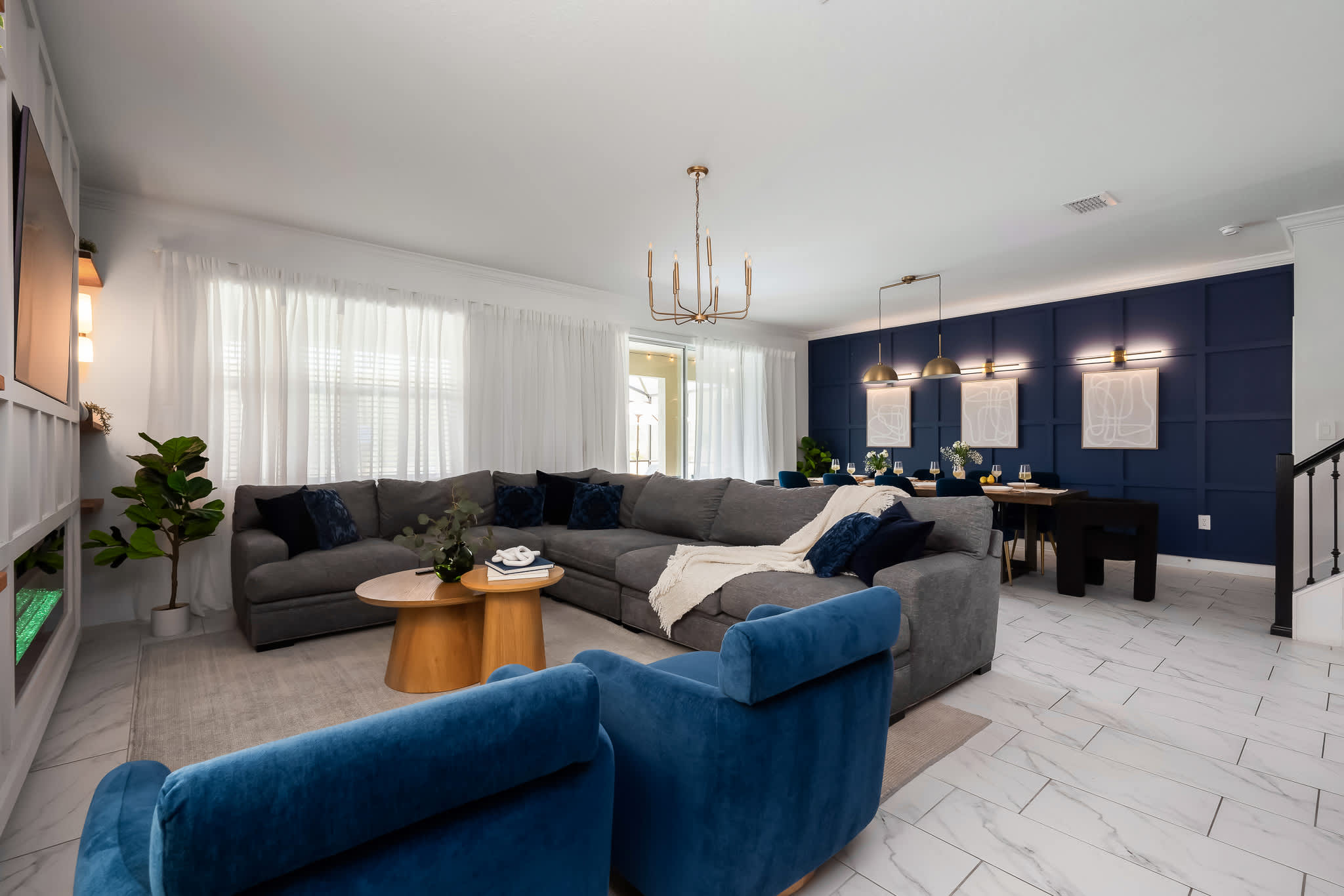 Open-concept living and dining area with a large gray sectional sofa, two blue armchairs, and a wooden coffee table. The dining area features a long wooden table with blue velvet chairs against a dark blue accent wall.