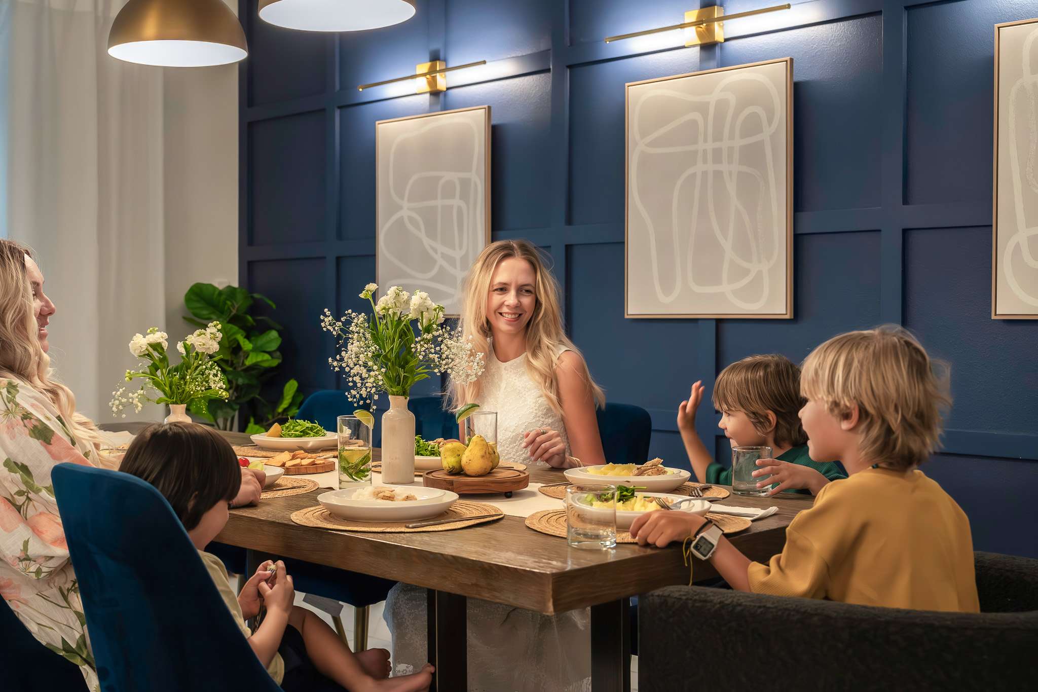 A modern dining room with a long wooden table set for a meal, surrounded by blue velvet chairs and a bench. The navy accent wall features decorative wainscoting and two abstract art pieces, illuminated by contemporary gold light fixtures.