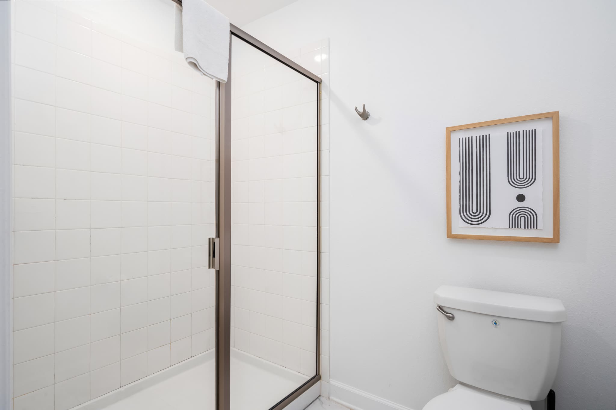 Bright white bathroom with a tiled walk-in shower featuring a bronze-framed glass door, adjacent to a white toilet and a framed abstract art piece on the wall.