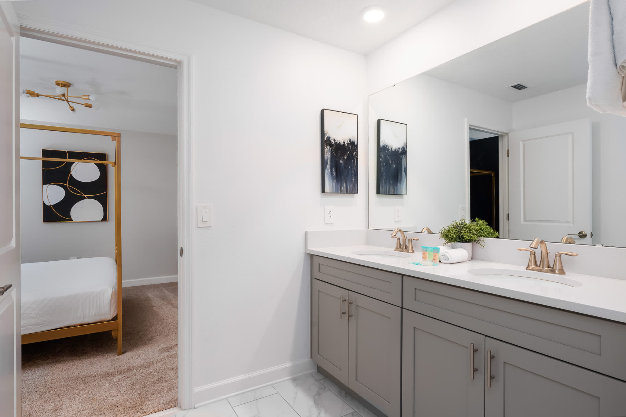 Modern bathroom featuring a double vanity with light gray cabinets, white countertops, and gold-toned fixtures, with a large mirror reflecting a doorway to a carpeted bedroom with a gold-framed bed.