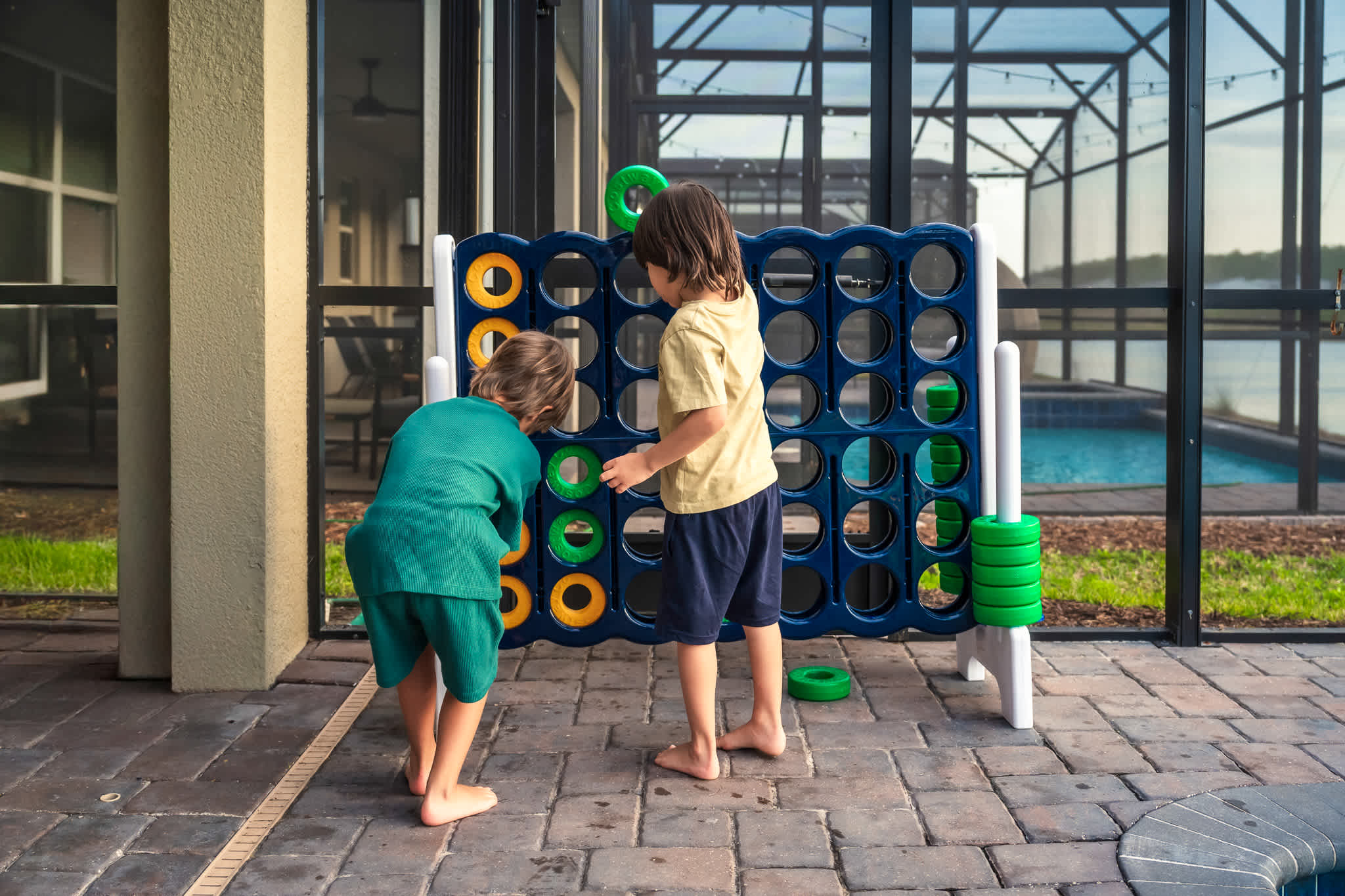Two children are engaged in playing a giant Connect Four game on the brick-paved patio, with the screened pool enclosure and a glimpse of the pool visible in the background.