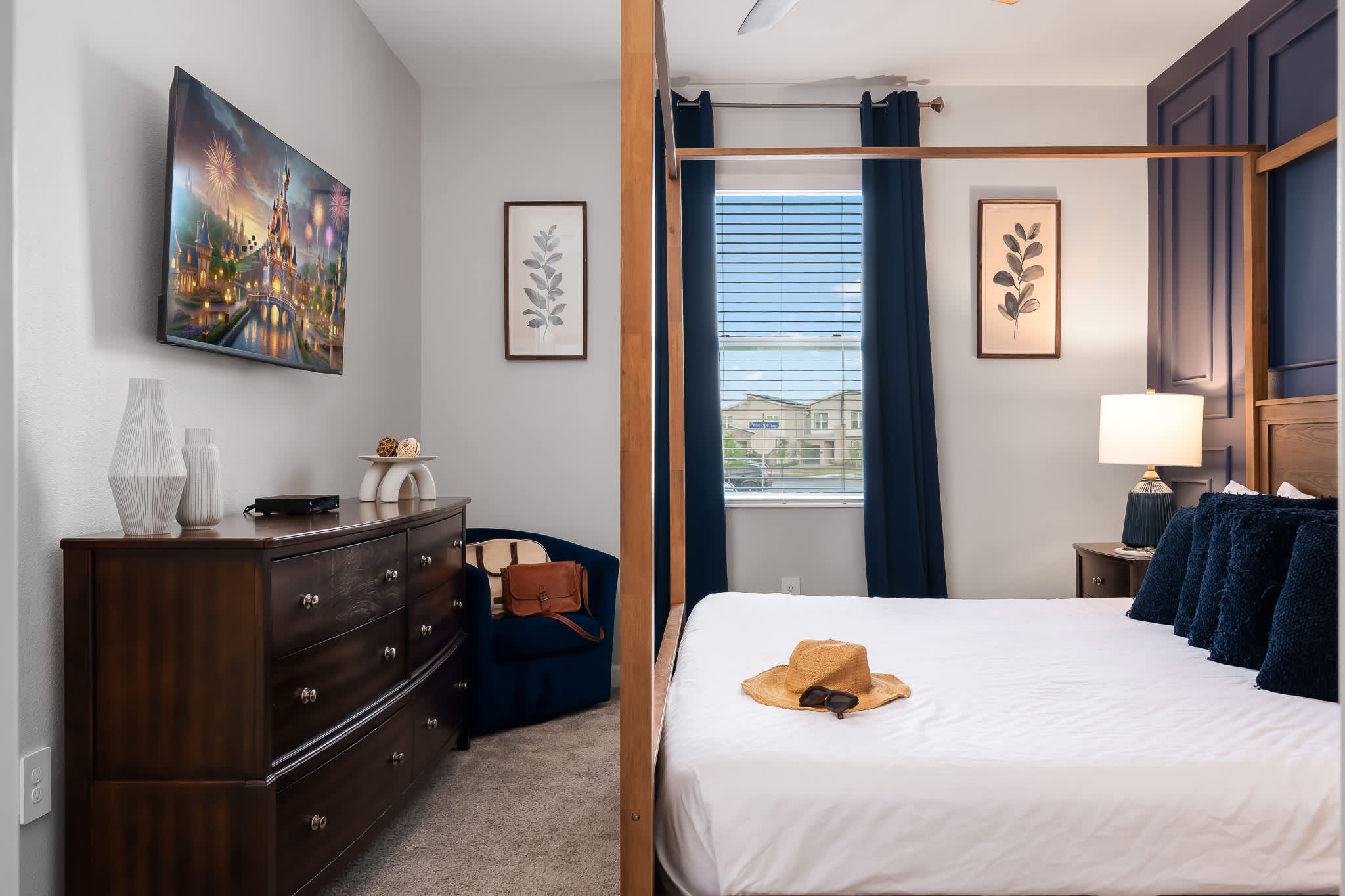 Bright bedroom featuring a wooden four-poster king bed, a dark wood dresser with a wall-mounted TV, and a window with blue blinds and curtains.