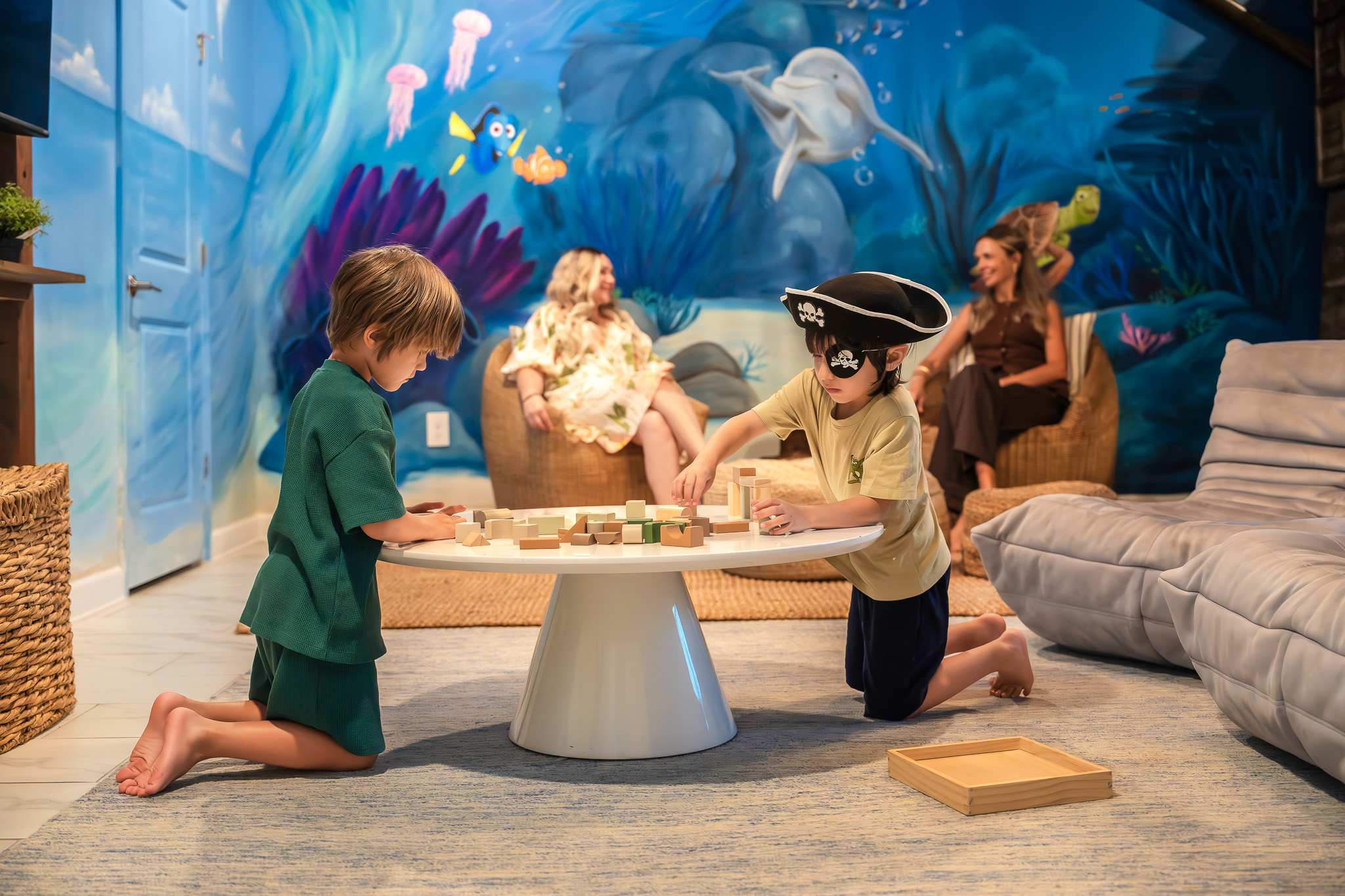 Two children play with wooden blocks on a white coffee table in a brightly painted ocean-themed room, while adults relax on woven chairs in the background. The walls are adorned with colorful underwater murals featuring marine life.