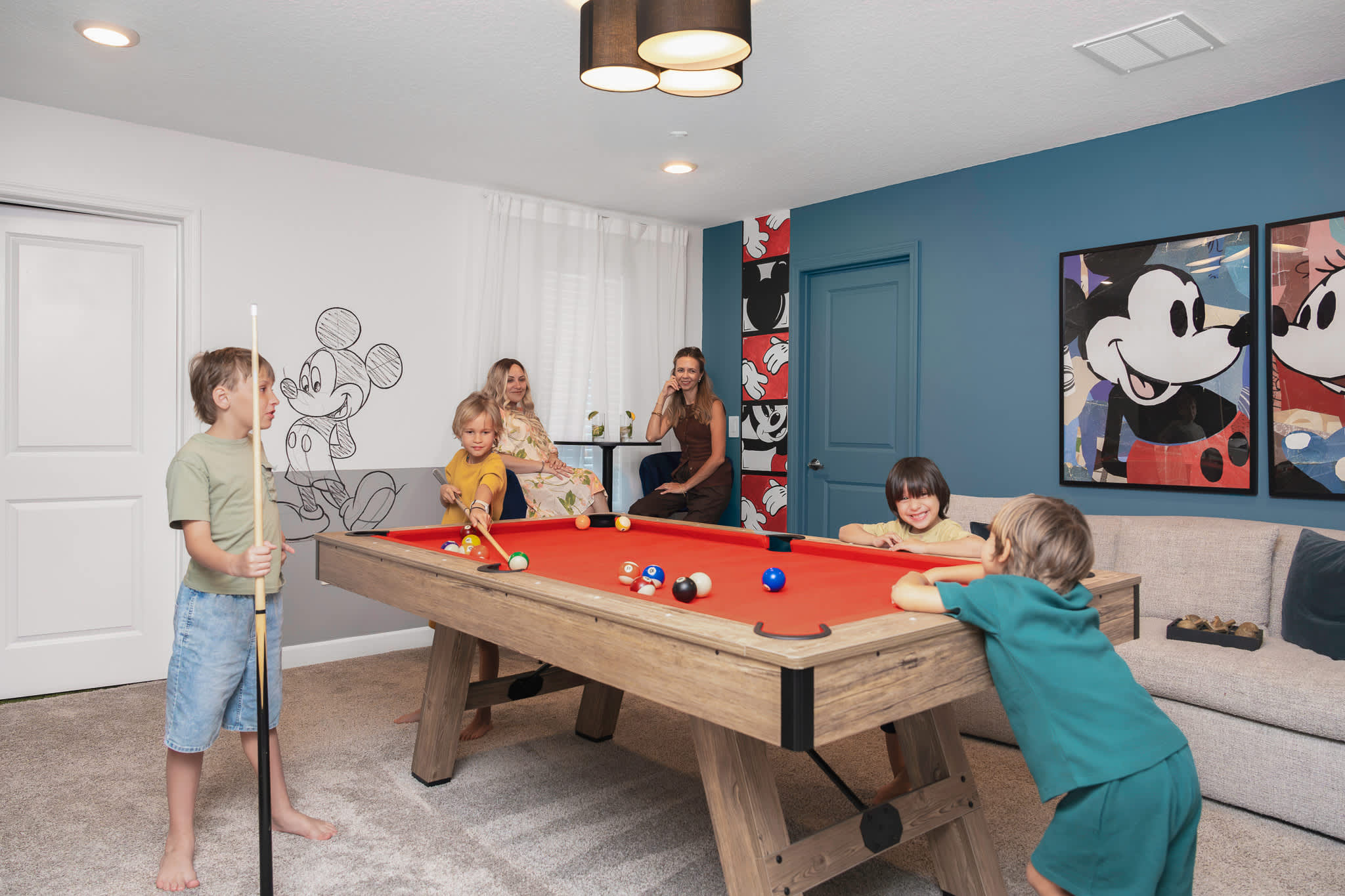 Children are actively playing pool on a red-felted pool table in a brightly lit game room, while two women relax at a high-top table in the background, surrounded by Mickey Mouse wall art.