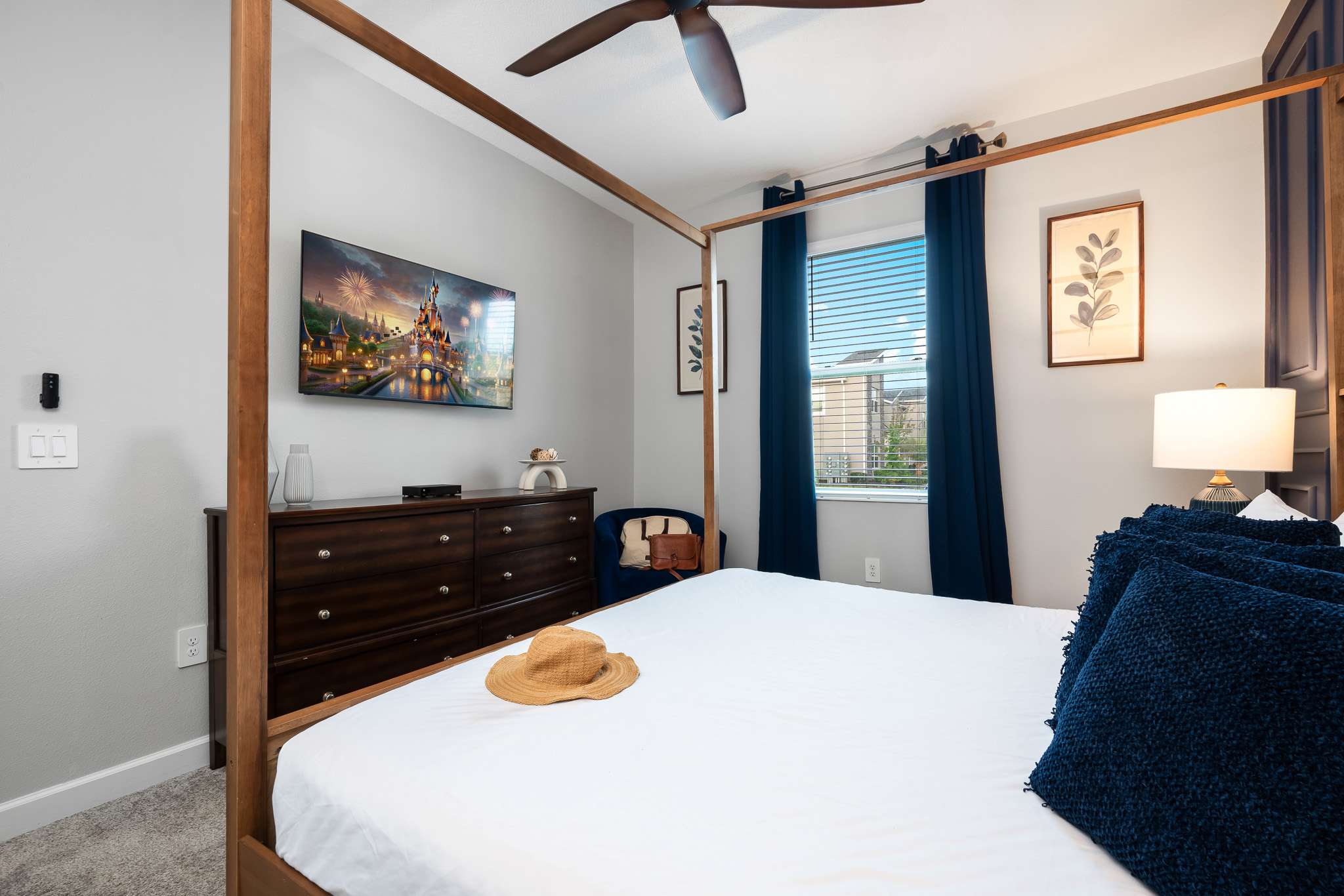 View of a bedroom with a wooden four-poster bed, a wall-mounted TV above a dark wood dresser, and a window with blue blinds and curtains.