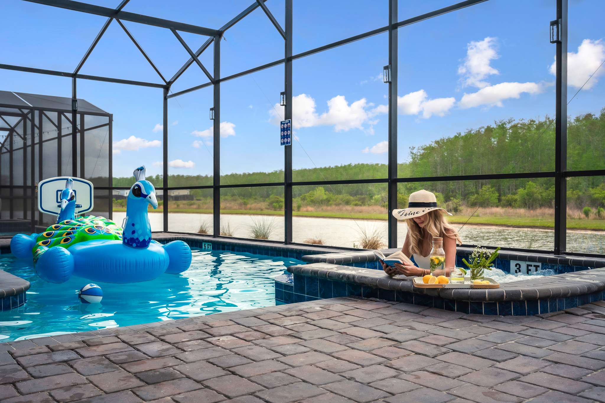 A woman in a straw hat reads a book while relaxing in the hot tub within a screened-in pool area, with a wooden tray holding drinks and fruit nearby, and inflatable peacock floats visible in the main pool.