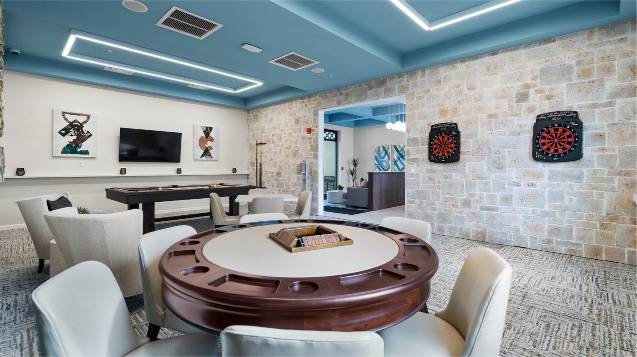 Vibrant game room featuring a dark wood poker table with cream chairs, a pool table in the background, and two dartboards mounted on a textured stone accent wall, all under a light blue ceiling with modern linear lighting.