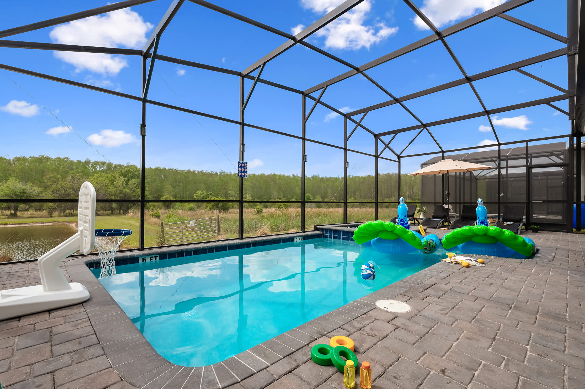 A wide shot of a screened-in private pool area with a basketball hoop and various pool floats, set on a paved deck. The enclosure offers expansive views of a natural, undeveloped landscape under a bright blue sky.