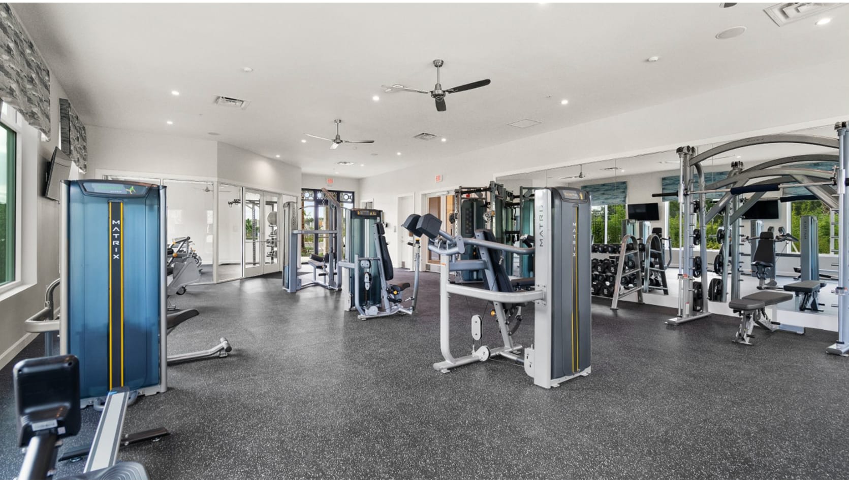 Well-equipped fitness center with a variety of modern exercise machines, including treadmills, ellipticals, and weight machines, set on a black rubber floor with large mirrors and windows offering natural light.