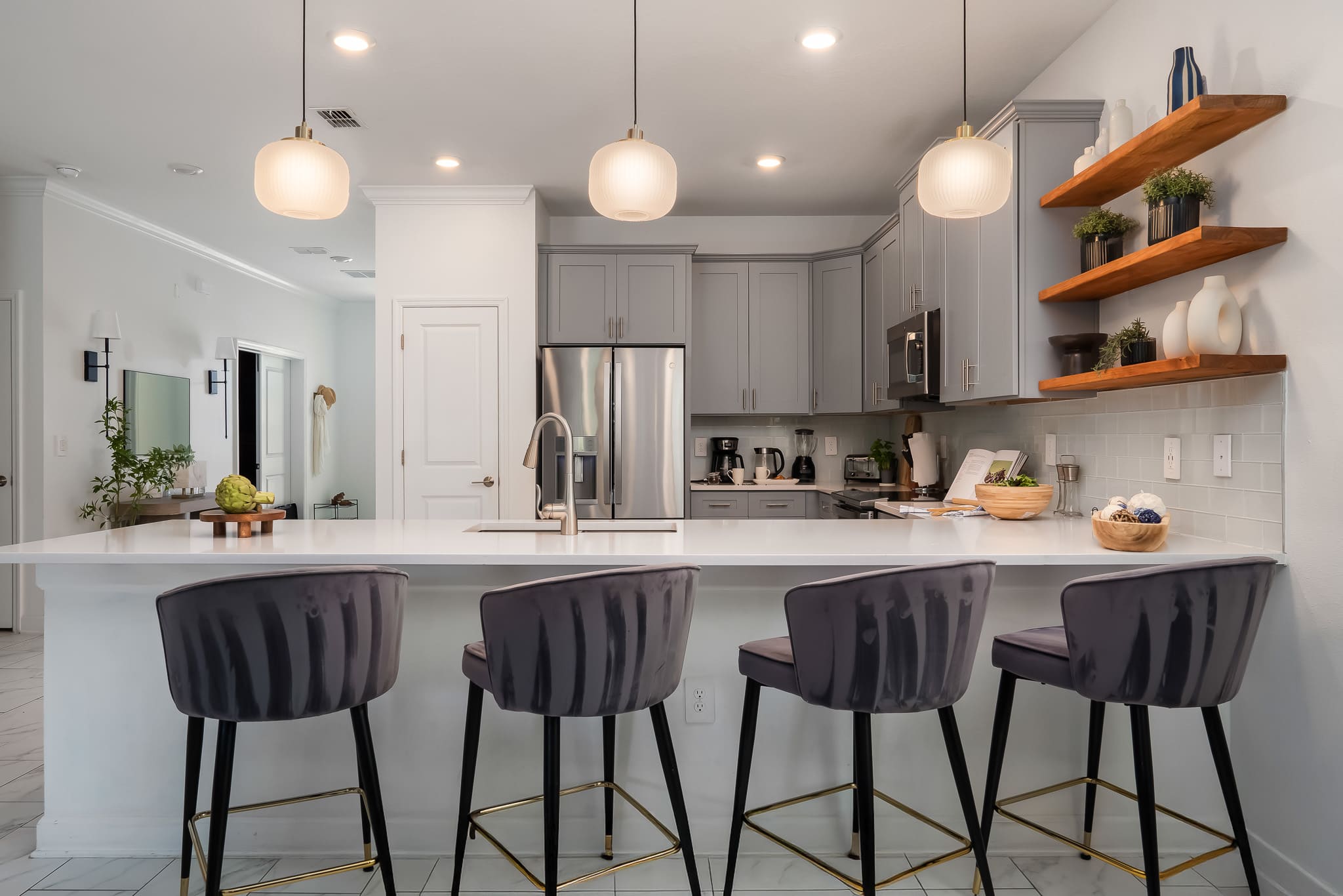 View of a modern kitchen from the dining area, showcasing a large white quartz island with four gray velvet bar stools, light gray shaker cabinets, and stainless steel appliances.
