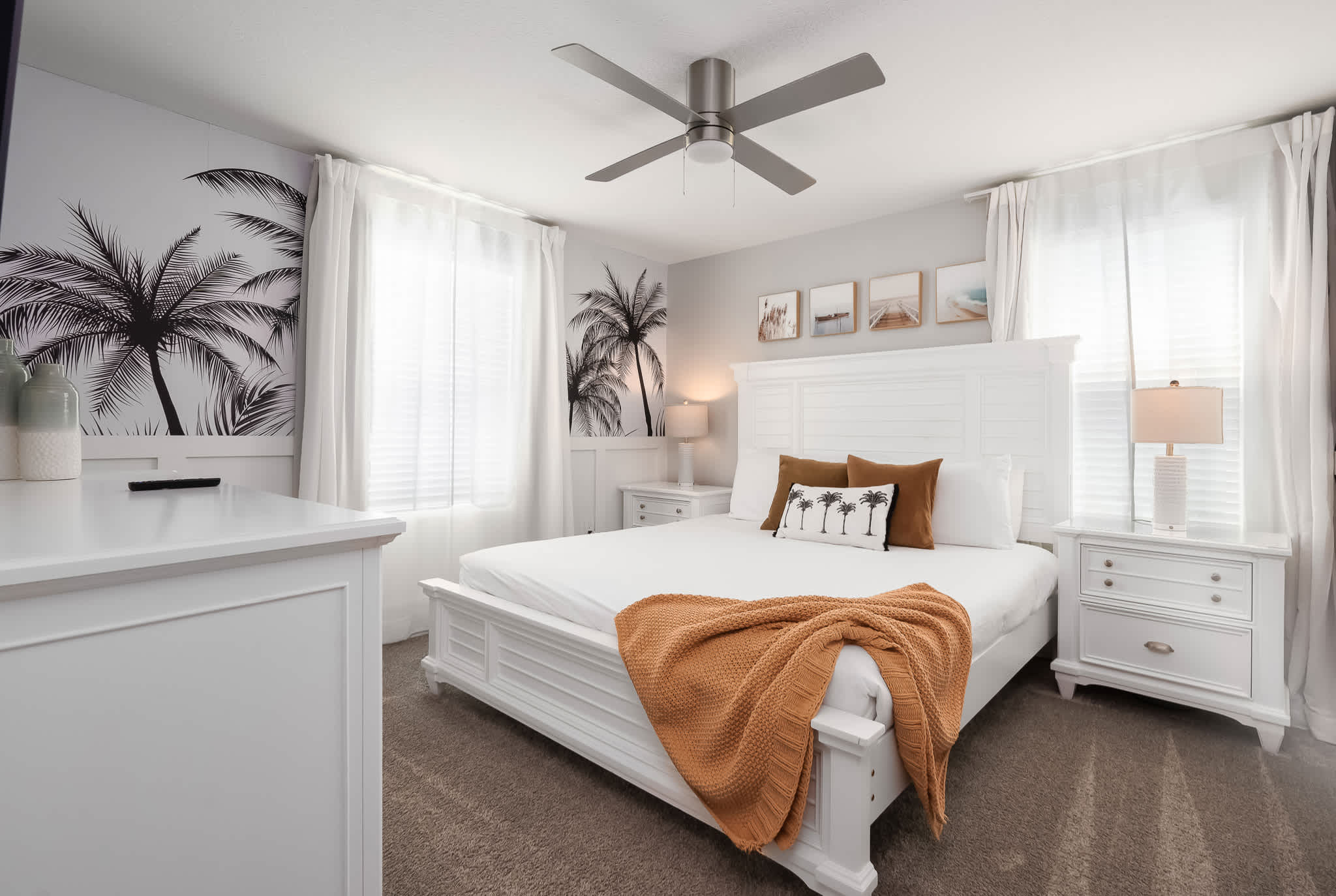 Serene tropical-themed bedroom featuring a white queen-sized bed with a tall headboard, palm tree wall decals, white dressers and nightstands, and a ceiling fan.