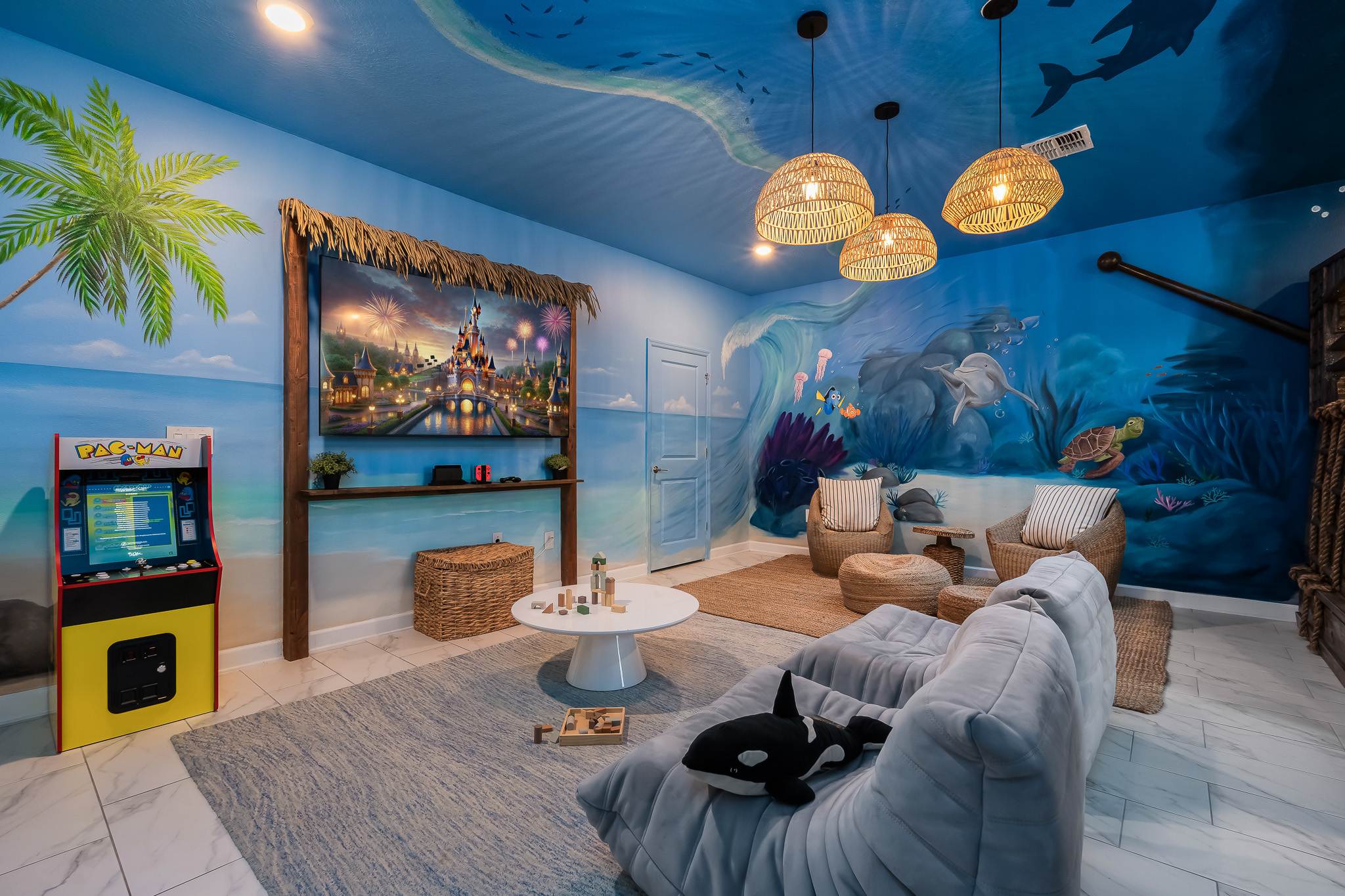 A lively ocean-themed game room features a large wall-mounted TV displaying a castle, a Pac-Man arcade machine, and comfortable gray bean bag seating. The walls and ceiling are painted with vibrant underwater and beach murals, illuminated by woven pendant lights.