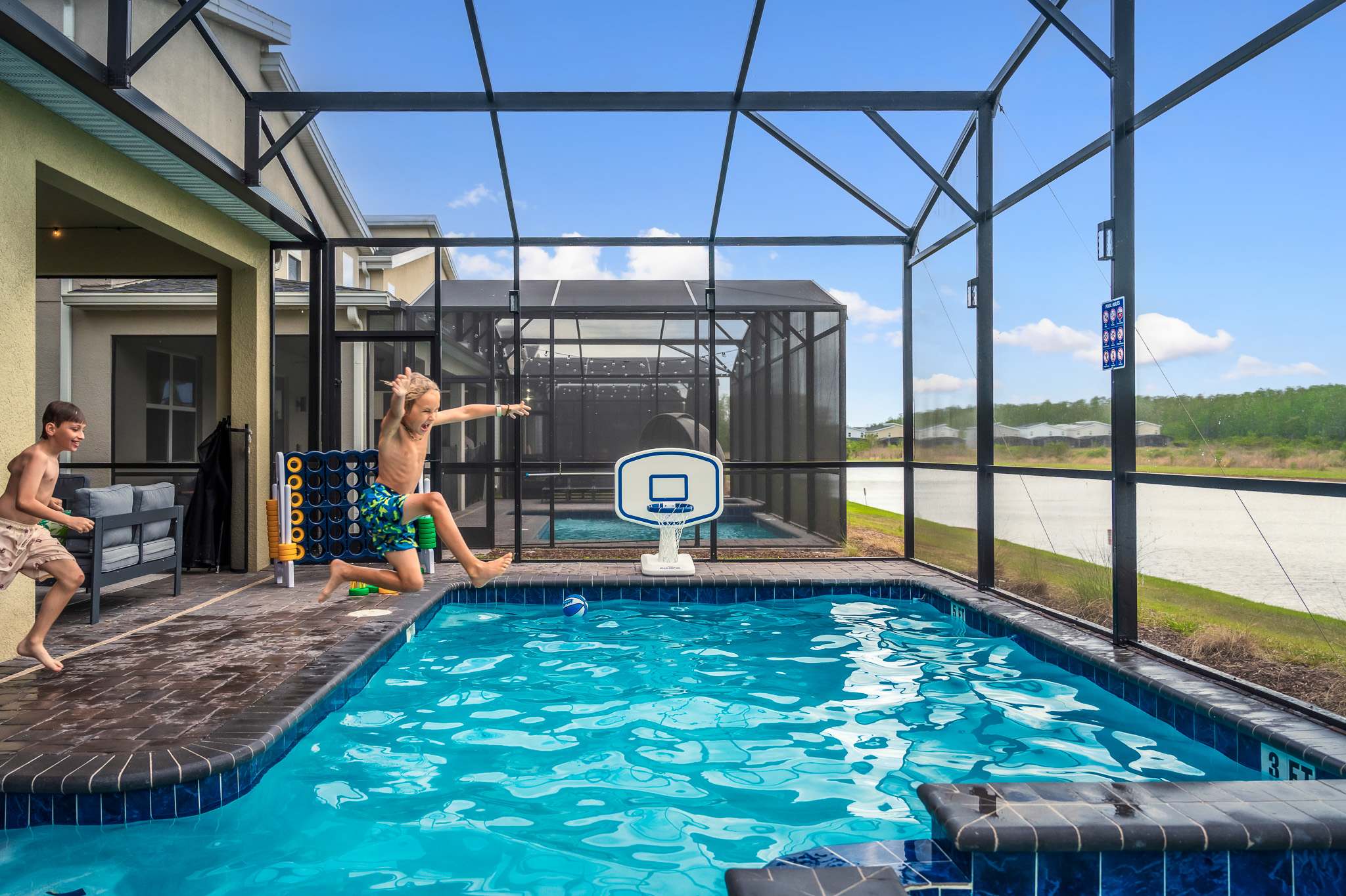 A child joyfully jumps into a screened-in private pool with a basketball hoop, surrounded by a paved deck and overlooking a natural landscape. The pool area is bright and open, with clear blue water.
