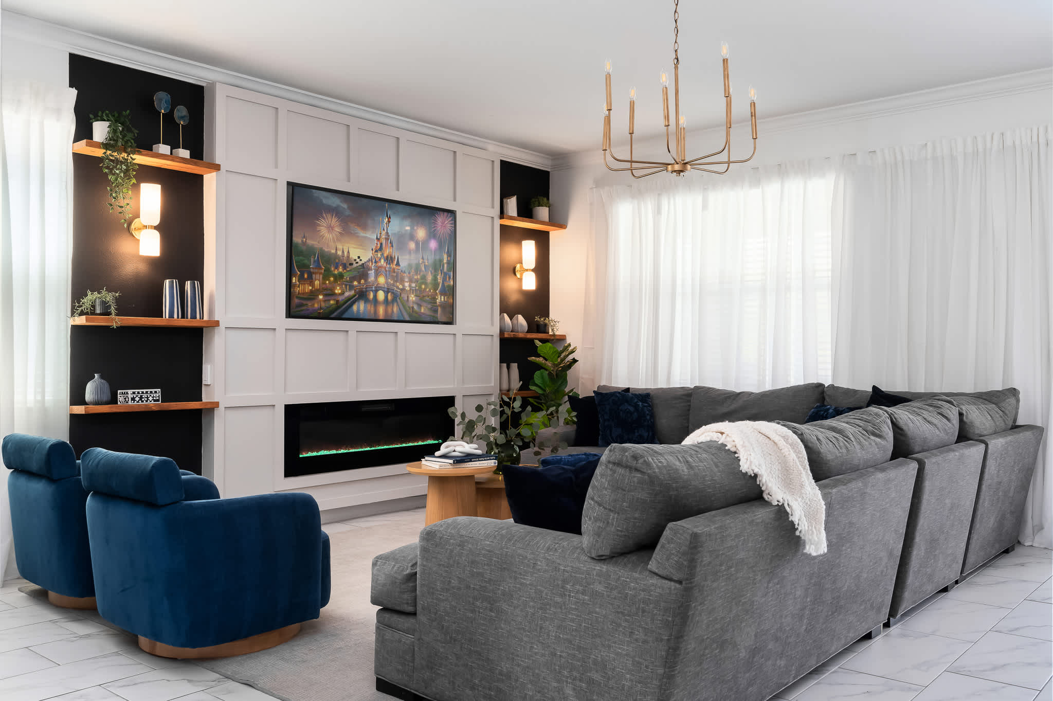 Spacious living room with a gray sectional sofa and two blue swivel armchairs, centered around a modern entertainment wall with a large TV and linear fireplace. The room is well-lit by natural light from large windows.