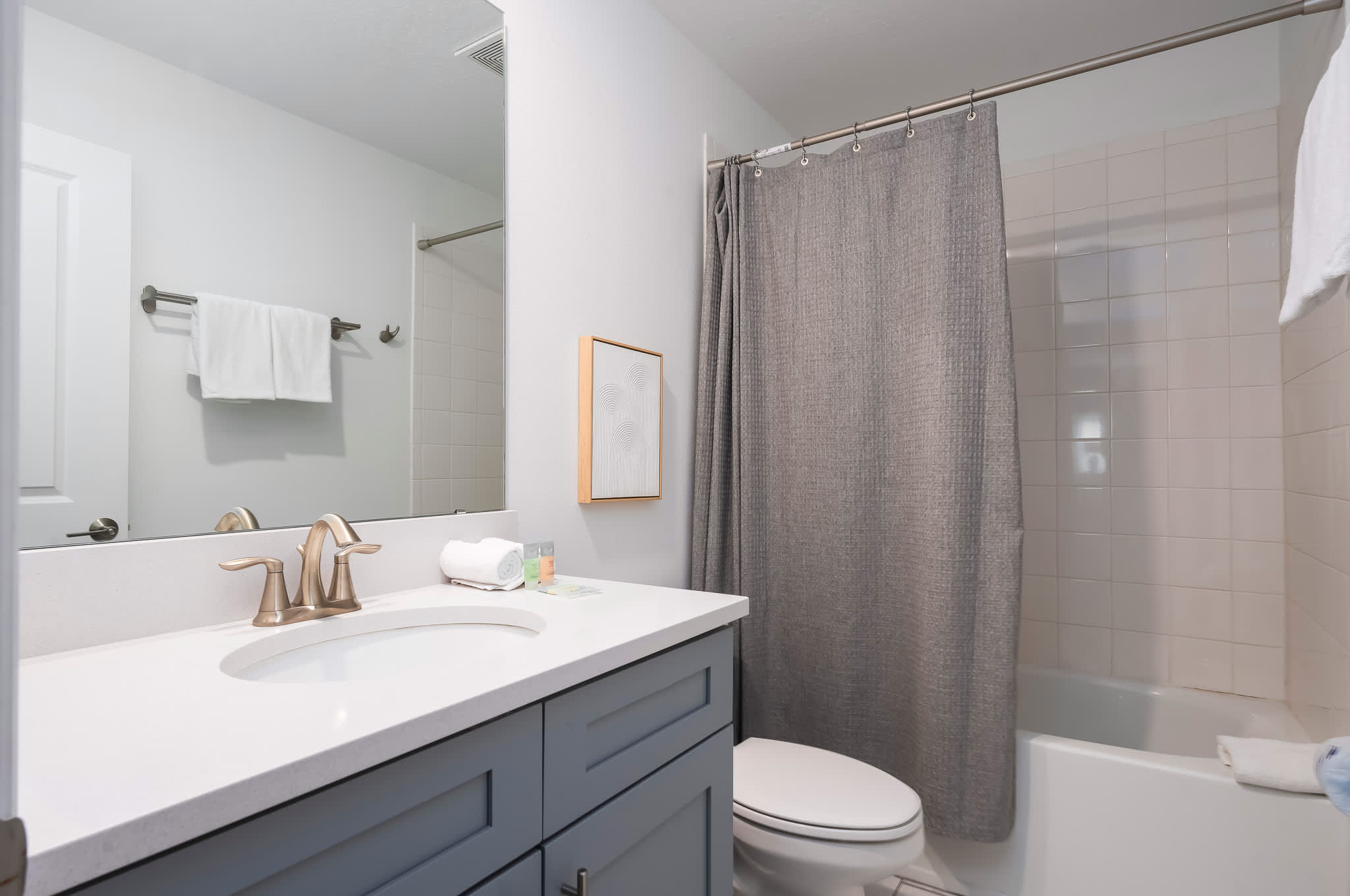 Clean and bright bathroom with a single gray vanity, white countertop, brushed gold faucet, a large mirror, and a bathtub with a shower curtain and white tiled walls.