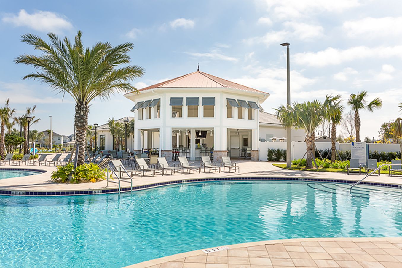 A large, elegant clubhouse building with a white facade and a distinctive octagonal copper-colored roof, situated beside a communal swimming pool with blue water and surrounding lounge chairs, all under a clear blue sky.