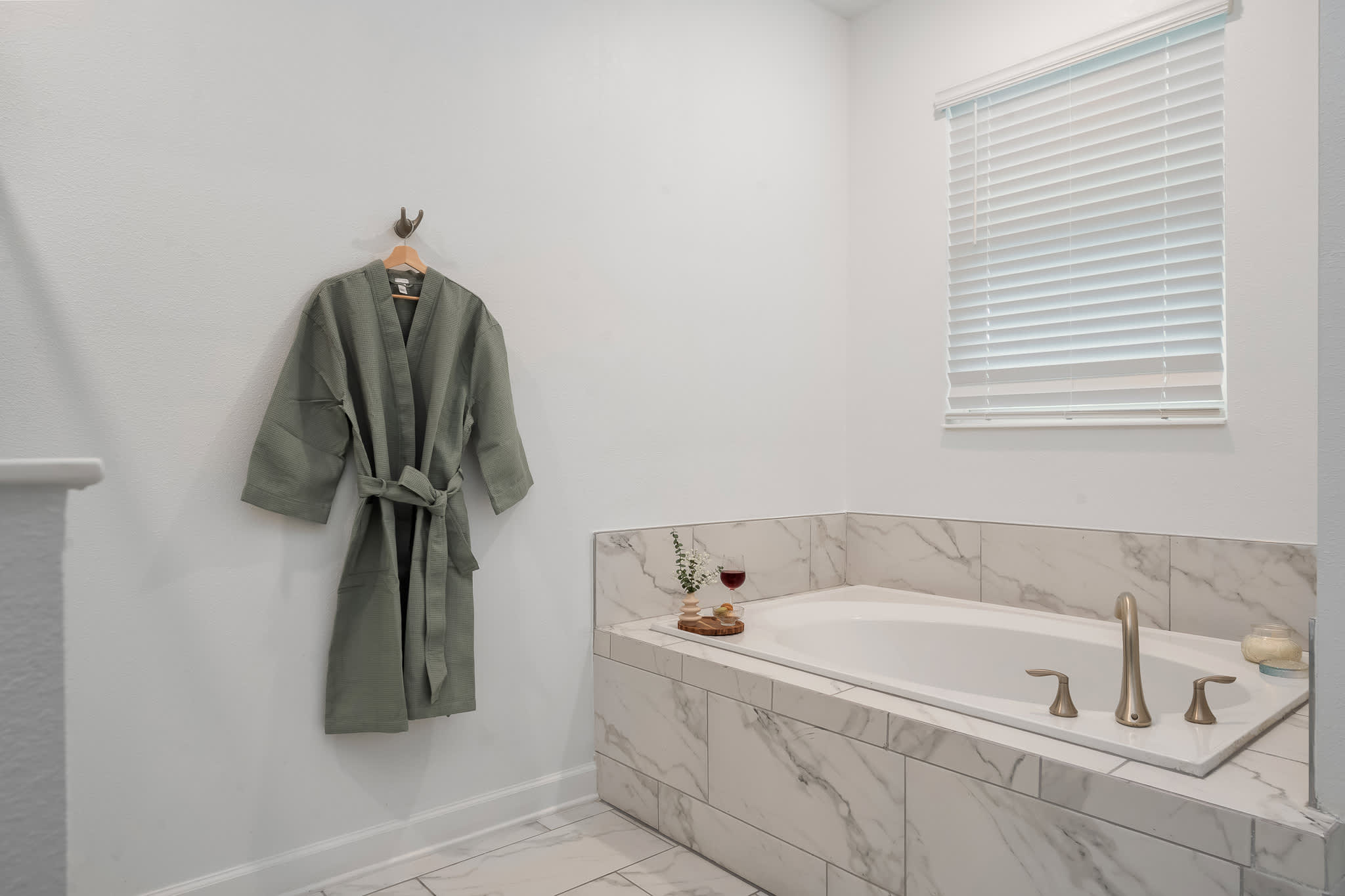 A bright white bathroom features a built-in bathtub with marble-look tile surround and gold fixtures, alongside a light green robe hanging on a wall hook. A window with white blinds is positioned above the tub.
