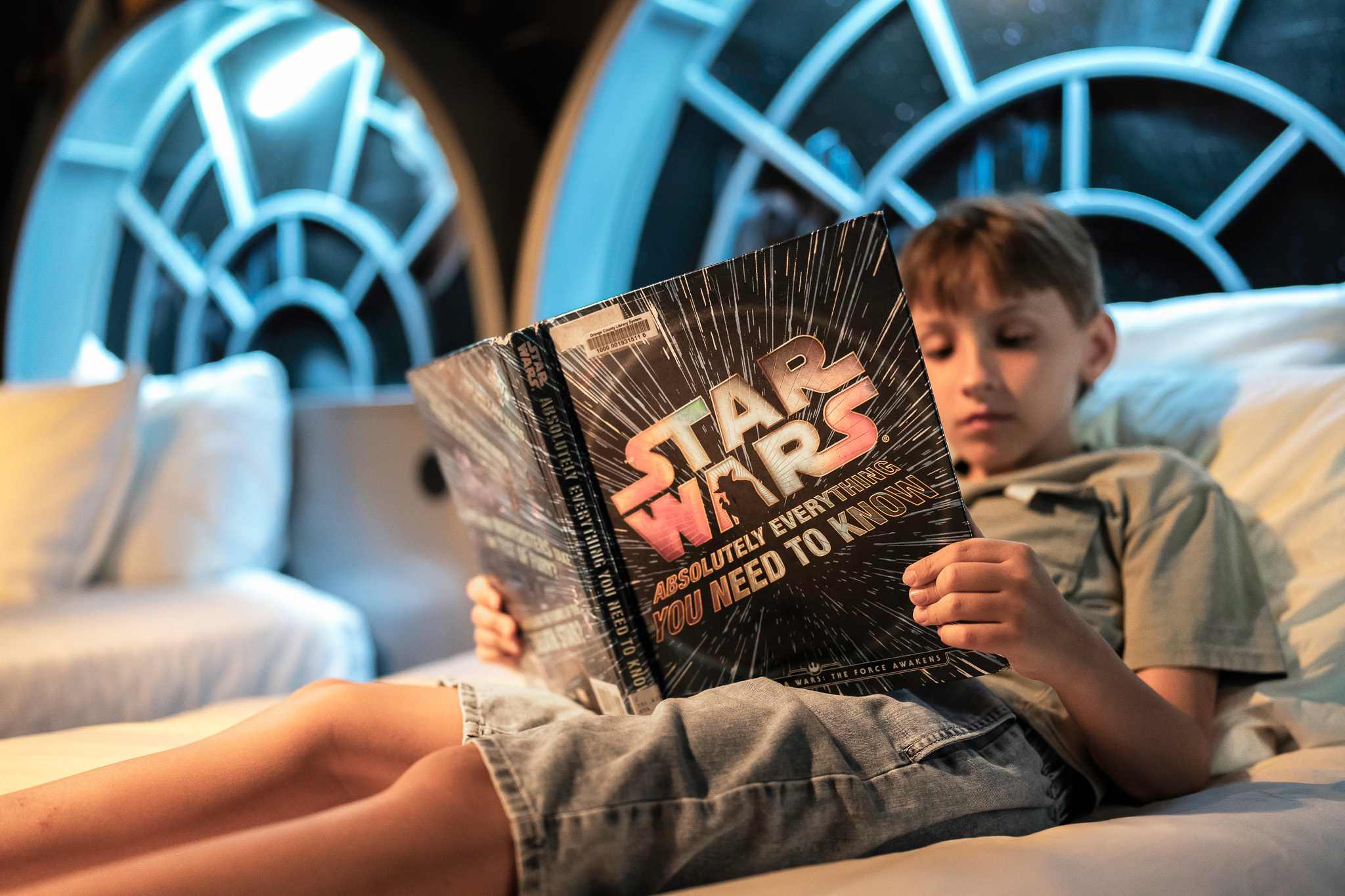 A child is comfortably reading a "Star Wars" book on one of the twin beds in the themed bedroom, which features an illuminated, circular spaceship window headboard and a vibrant space-themed wall mural.