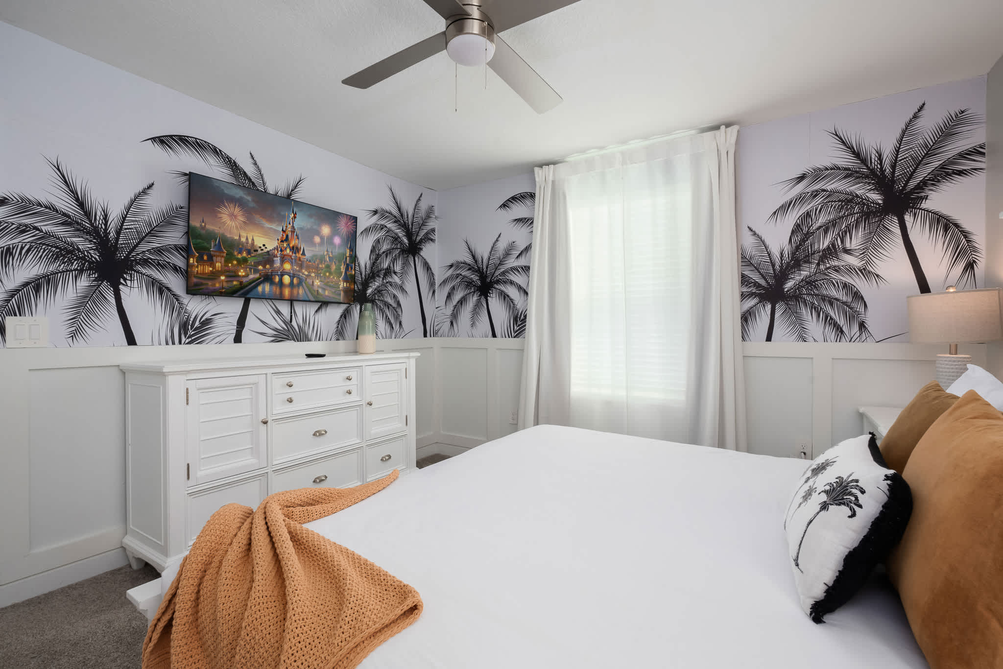 A tropical-themed bedroom featuring a white dresser with a TV displaying a castle scene, set against a white wall with black palm tree silhouettes, and a bed with white bedding and an orange throw.