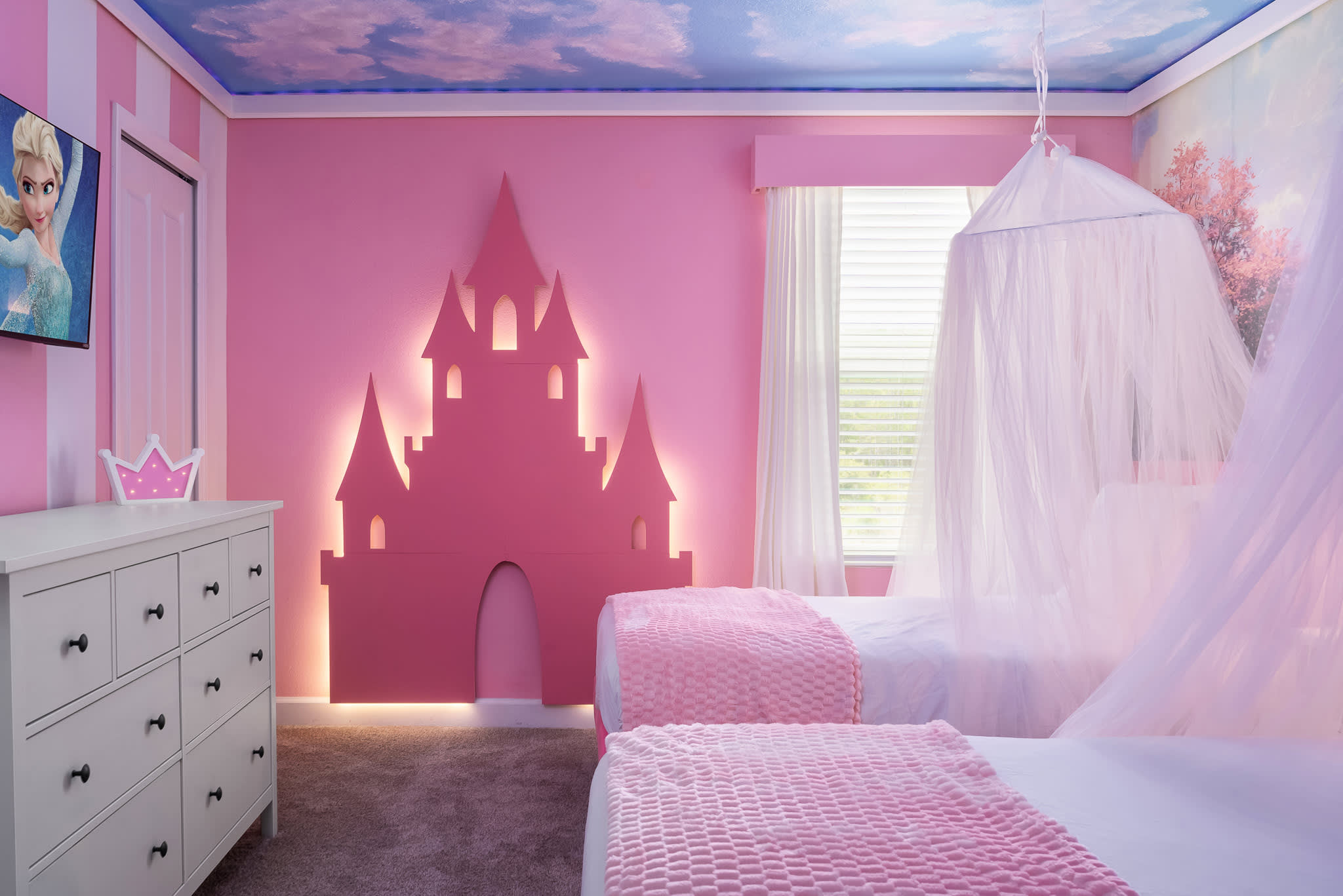 Bright princess-themed bedroom with two pink twin beds, sheer canopies, a large pink castle wall decor with ambient lighting, and a window with white blinds and curtains.