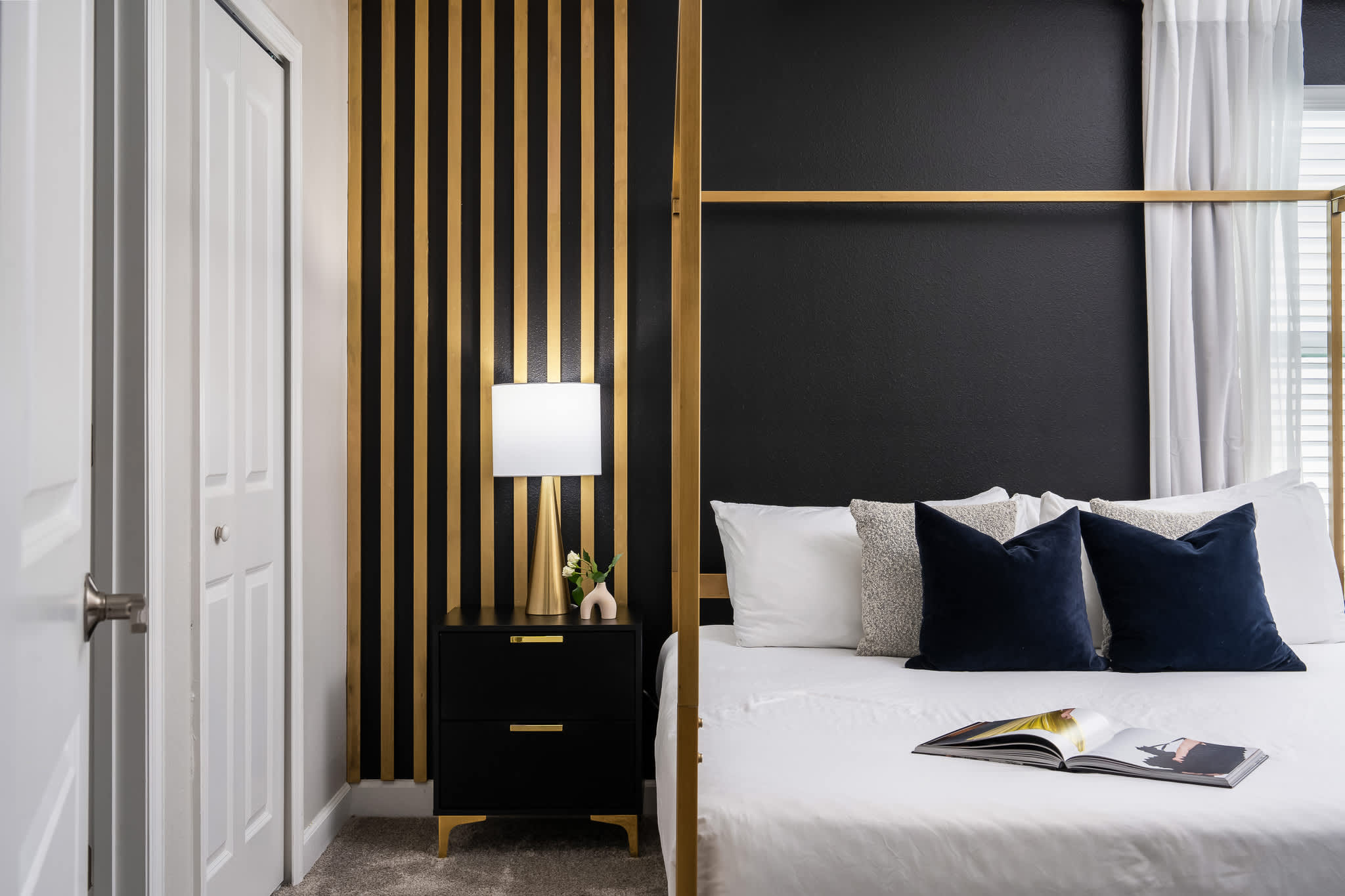 Close-up of the elegant bedroom showcasing a black nightstand with a gold lamp, next to a gold four-poster king bed, against a black accent wall with vertical gold slats, and a white closet door.