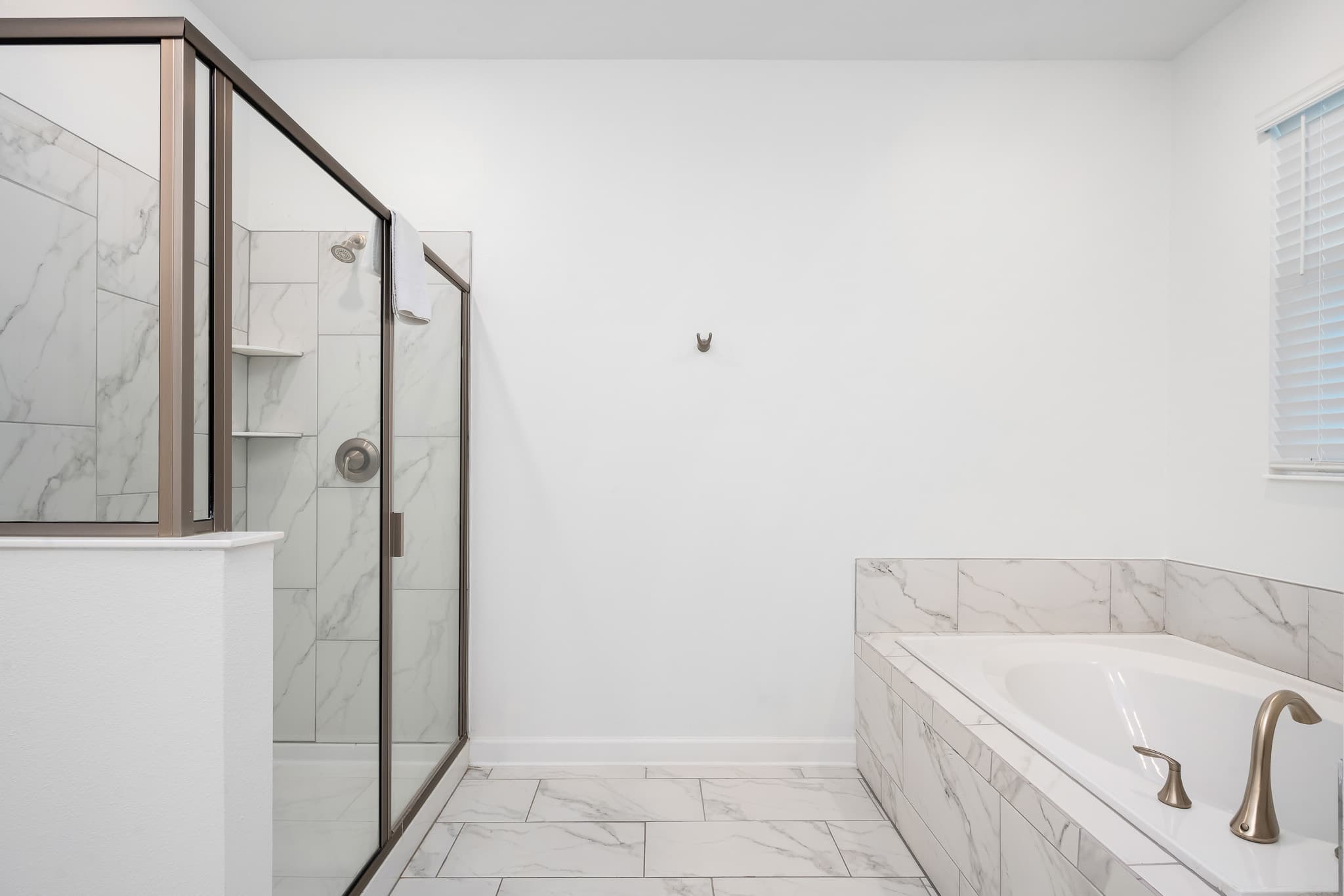 Modern bathroom featuring a spacious walk-in shower with glass enclosure and white marble-look tile, alongside a built-in bathtub with gold fixtures, all set against white walls and matching floor tiles.