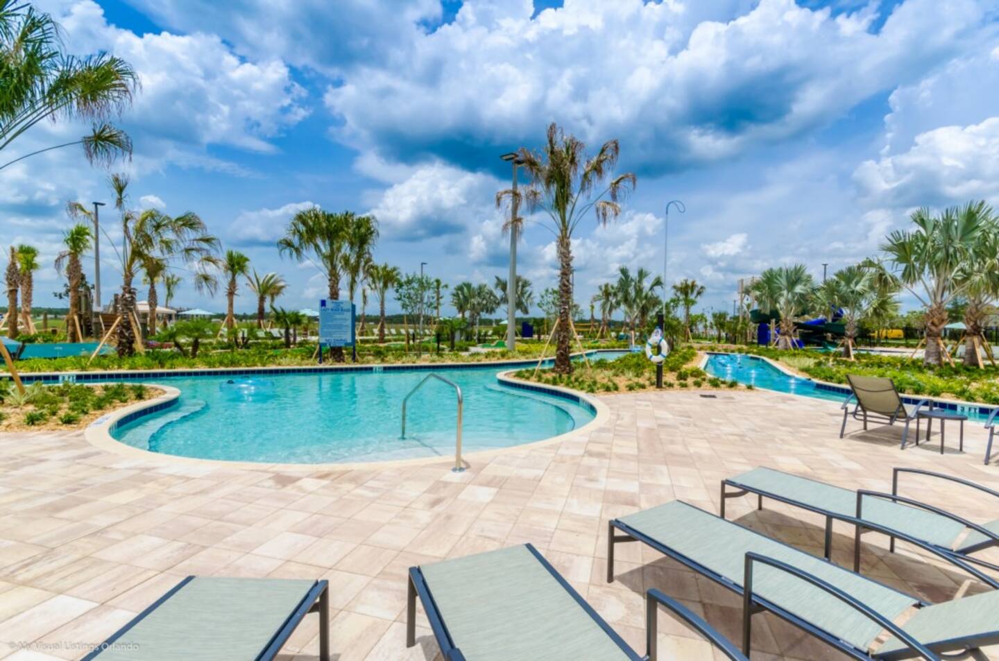 Sunny view of a communal swimming pool with light blue water, surrounded by light-colored paver decking and several green lounge chairs, with palm trees and a distant water park feature under a bright blue sky.