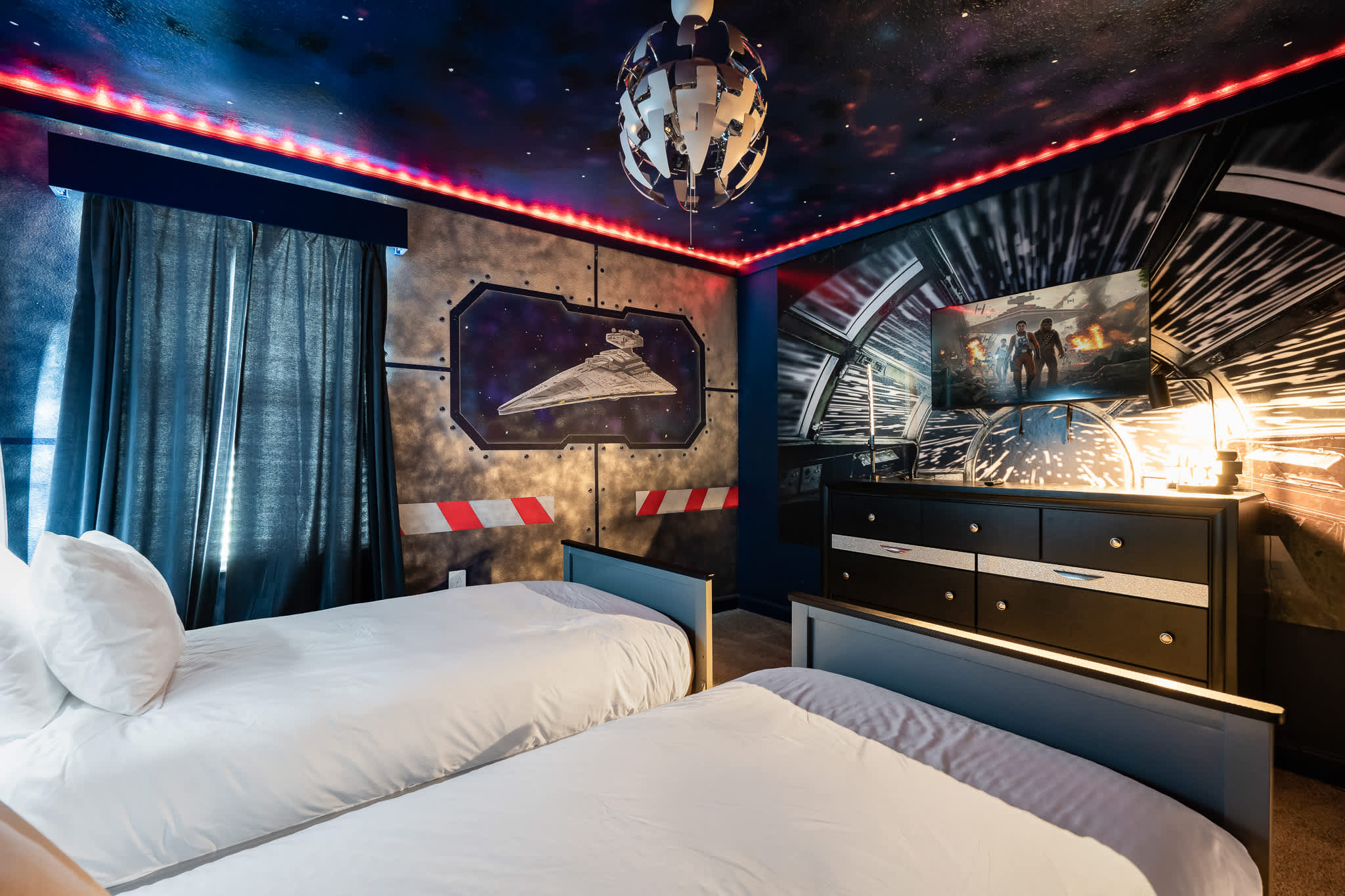 Themed bedroom with two twin beds, a dark dresser with a TV displaying a space scene, and a wall mural featuring a Star Destroyer, all under a starry ceiling with red accent lighting.