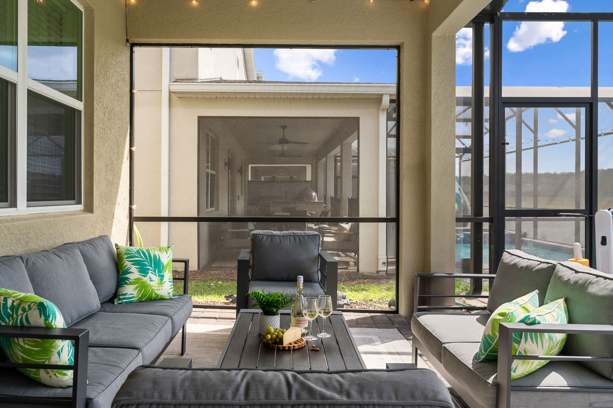 A comfortable outdoor patio area with grey wicker sofas and armchairs, adorned with green tropical pillows, centered around a coffee table with snacks and drinks. The screened enclosure provides a view of another screened pool area and the house exterior.