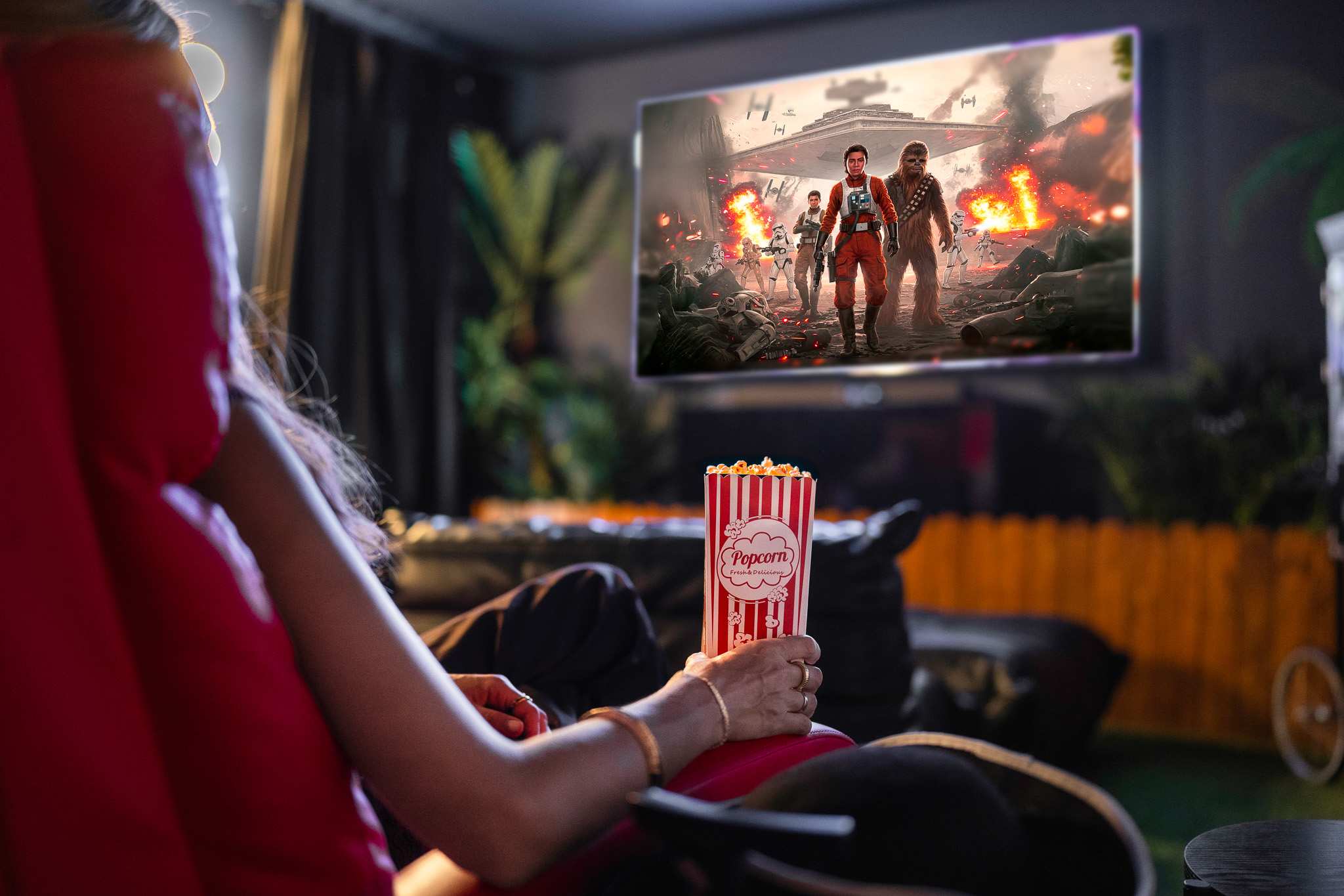 A person holds a box of popcorn while watching a Star Wars-themed movie on a large screen in a dimly lit media room, seated in a comfortable black chair.