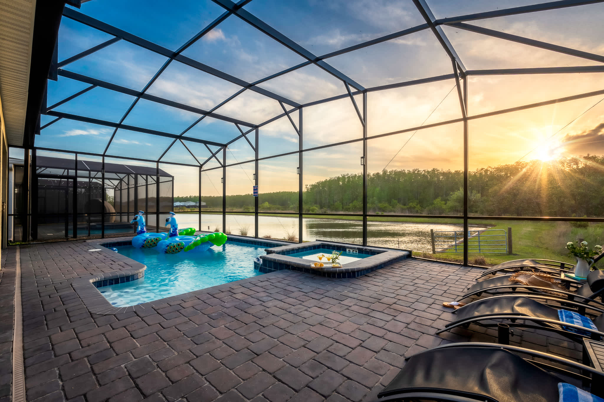 Screened-in outdoor pool and hot tub area with dark paver patio, several lounge chairs, and a scenic view of a lake and forest at sunset.