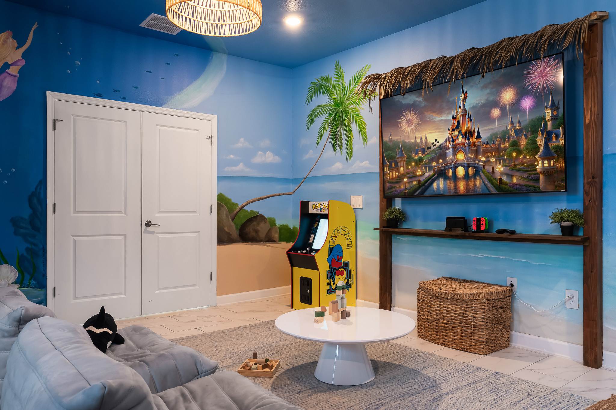 A corner of the ocean-themed game room showcases a large wall-mounted TV under a thatched canopy, a yellow Pac-Man arcade machine, and a white coffee table with wooden blocks. The walls are painted with a beach scene, and a plush orca toy rests on a gray sofa.