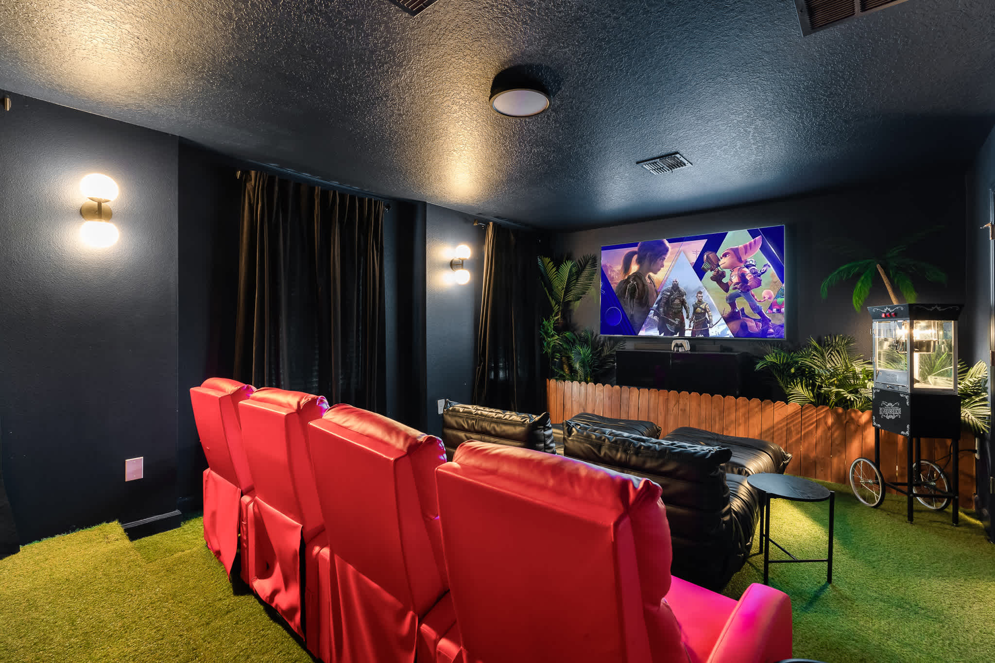 A dark and immersive media room with rows of red and black reclining theater seats facing a large screen displaying a video game. The room features dark walls, black curtains, and artificial turf flooring, with tropical plant decor and wall sconces.