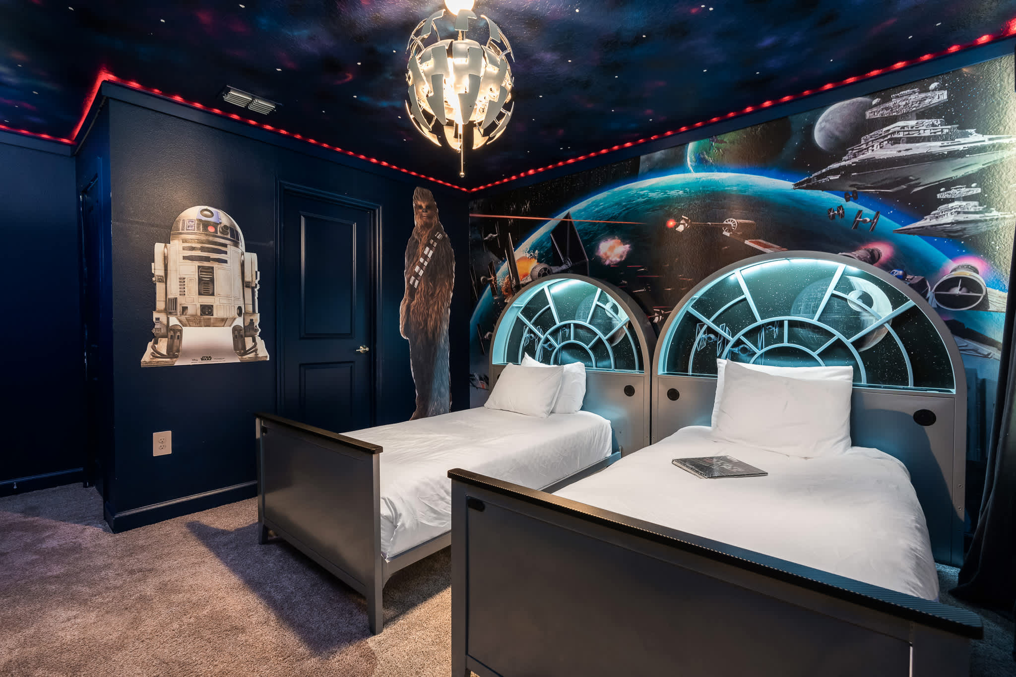 Themed bedroom with two twin beds featuring illuminated, circular headboards resembling spaceship windows, set against a dark blue wall mural depicting a space battle and a starry ceiling with red accent lighting.