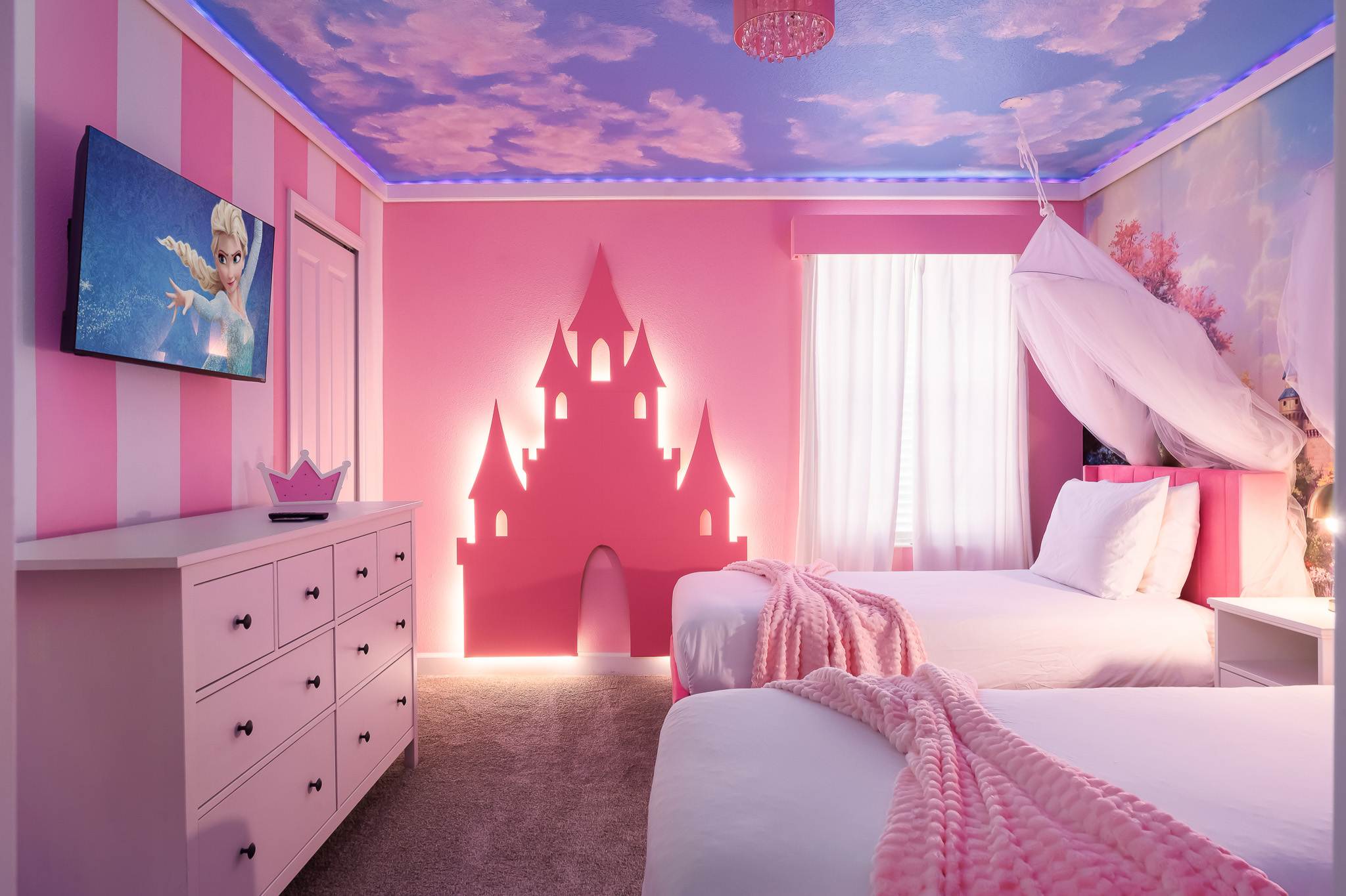 Enchanting princess-themed bedroom featuring two pink twin beds, a large pink castle headboard with backlighting, a pink dresser with a TV displaying a cartoon, and a cloud-painted ceiling with LED lights.