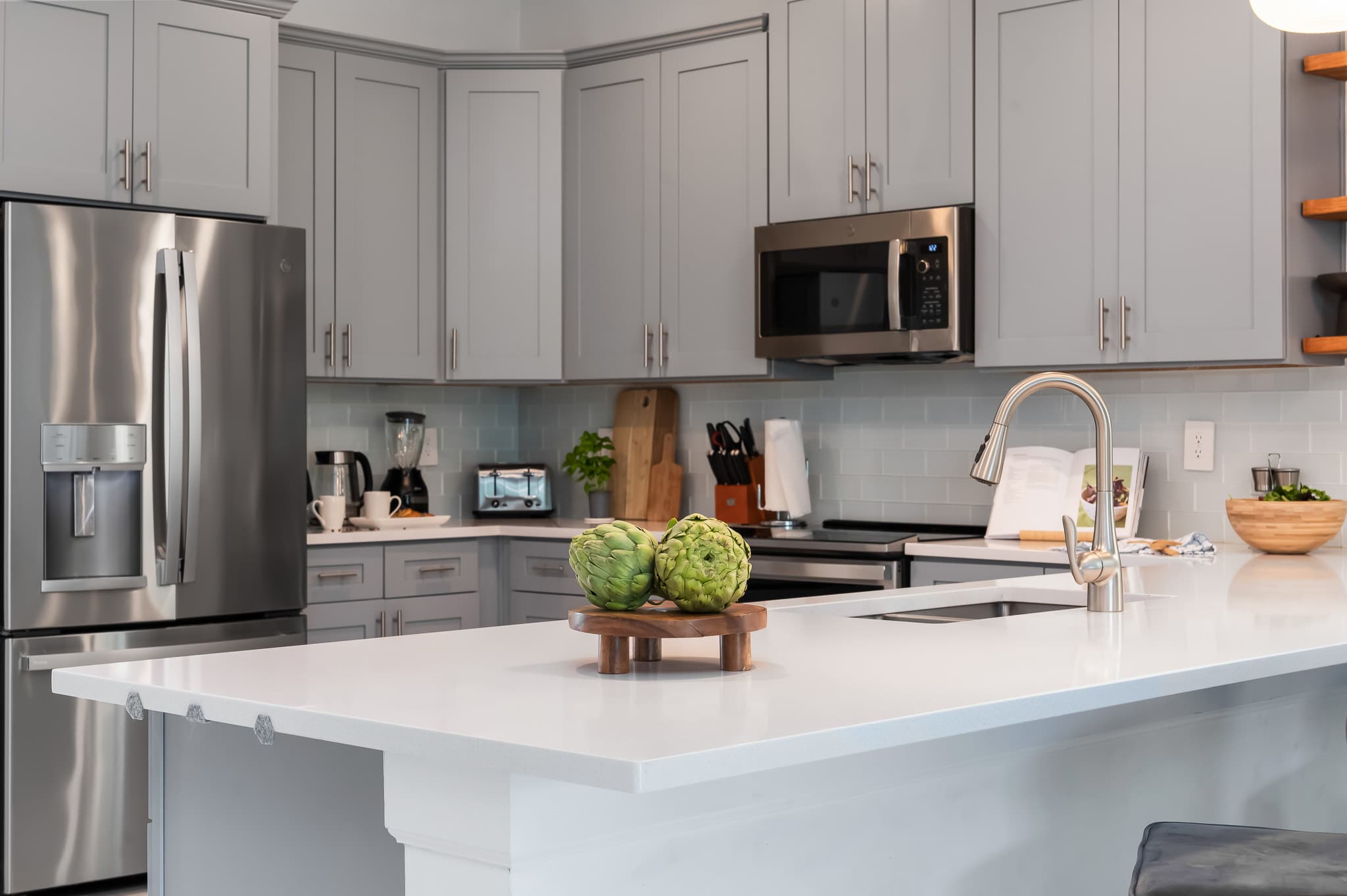 Close-up of a modern kitchen island with white quartz countertops, featuring decorative green artichokes on a wooden tray, and a stainless steel sink with a gooseneck faucet.